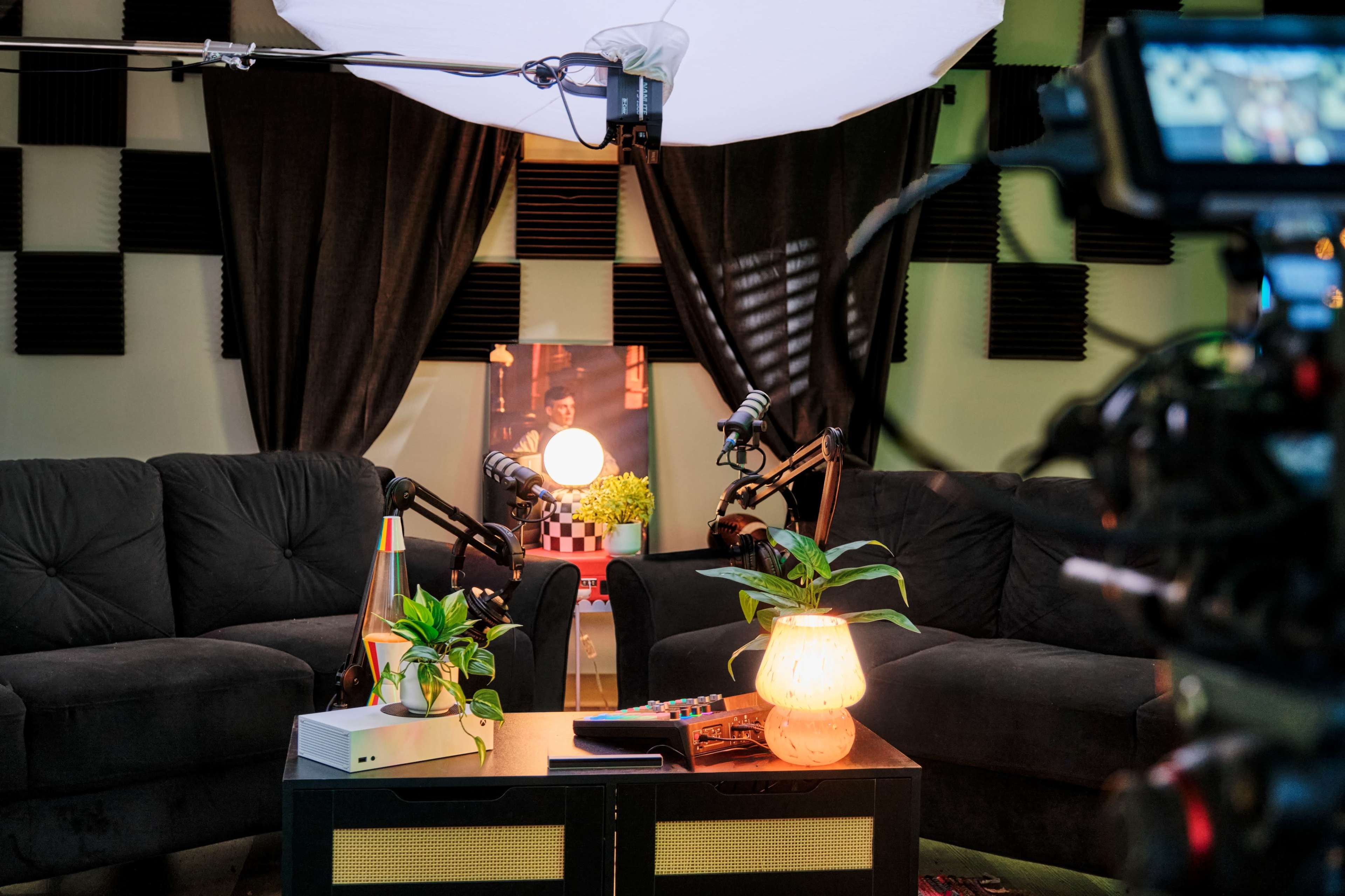 A cozy recording setup with two dark sofas, a coffee table adorned with a plant and decorative items, and a backdrop featuring curtains and soundproof panels.