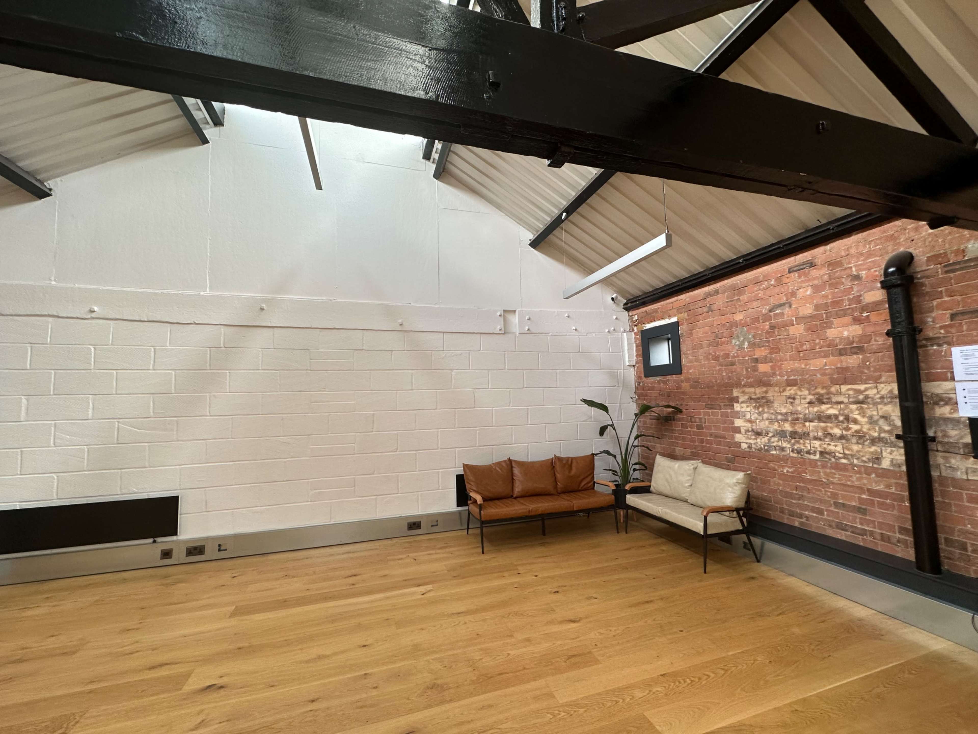 A spacious room with a wooden floor, featuring a leather sofa and a chair against a brick wall, under a high ceiling with exposed beams.