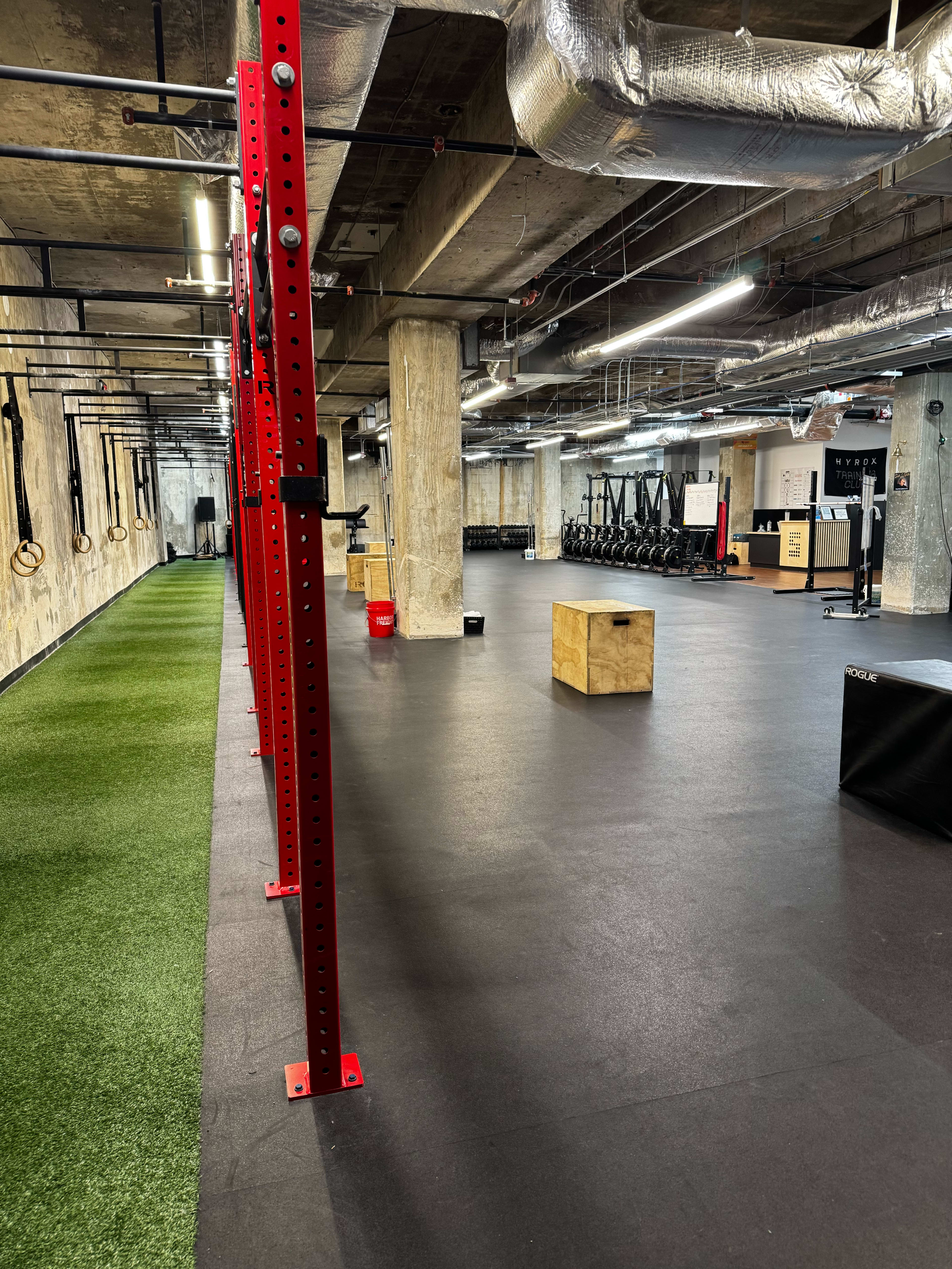 The image shows a spacious gym interior with a red pull-up rig on the left, rubber flooring, and various exercise equipment arranged in the background.