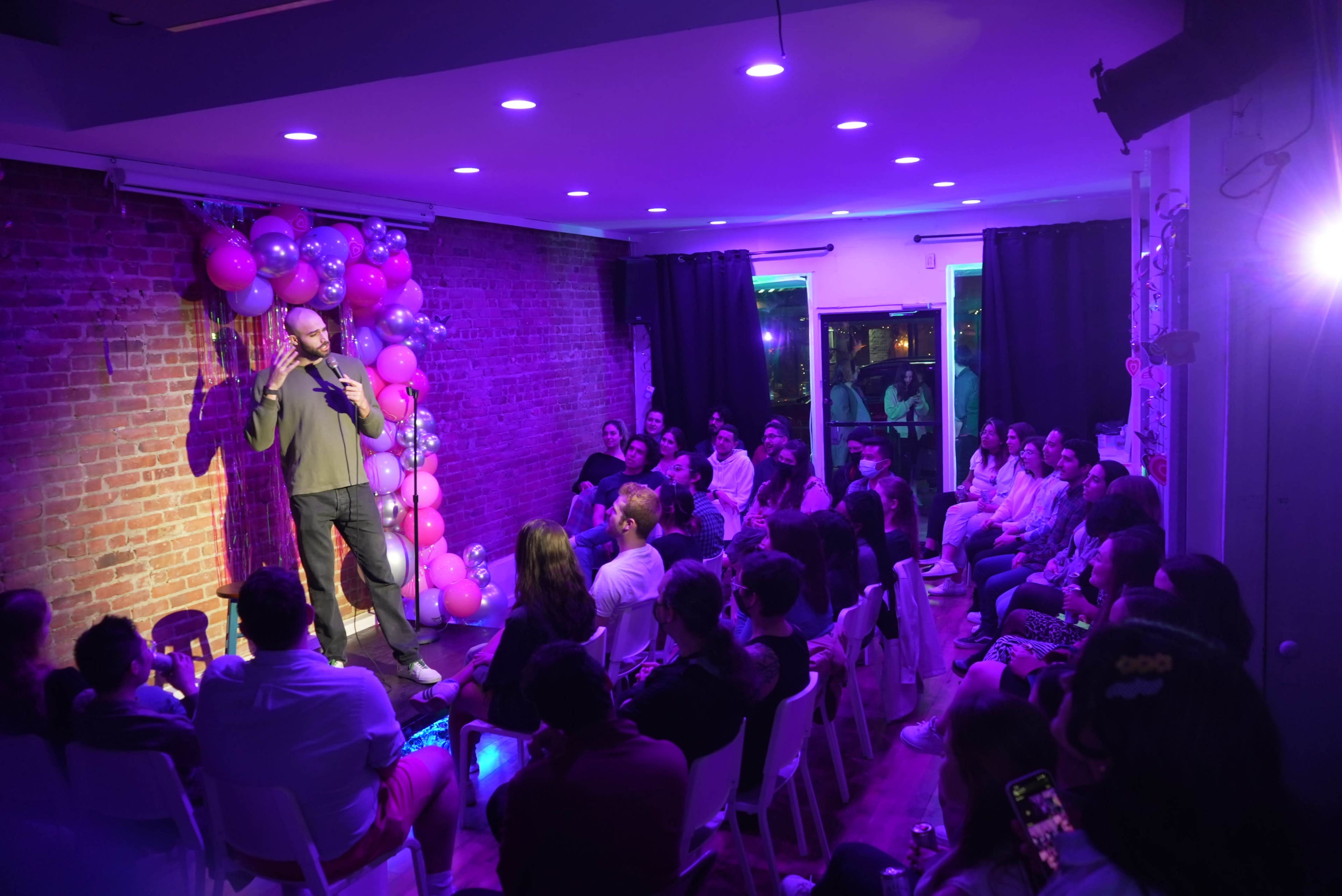 A comedian performs on stage in front of an audience seated in a brightly lit room decorated with pink balloons and exposed brick walls.