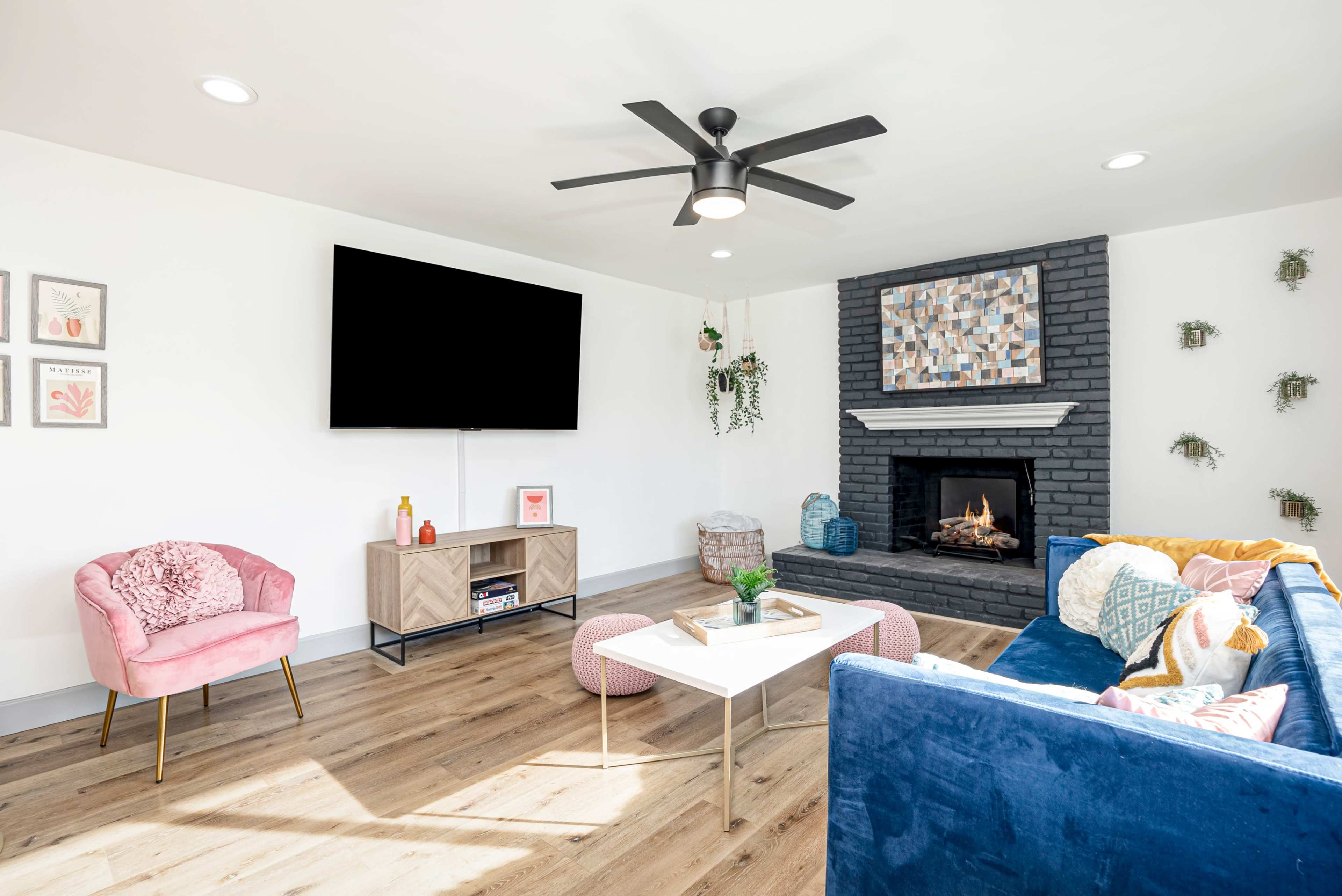 A modern living room features a blue couch, a white coffee table, a pink accent chair, a wall-mounted TV, and a fireplace with a decorative mantel.