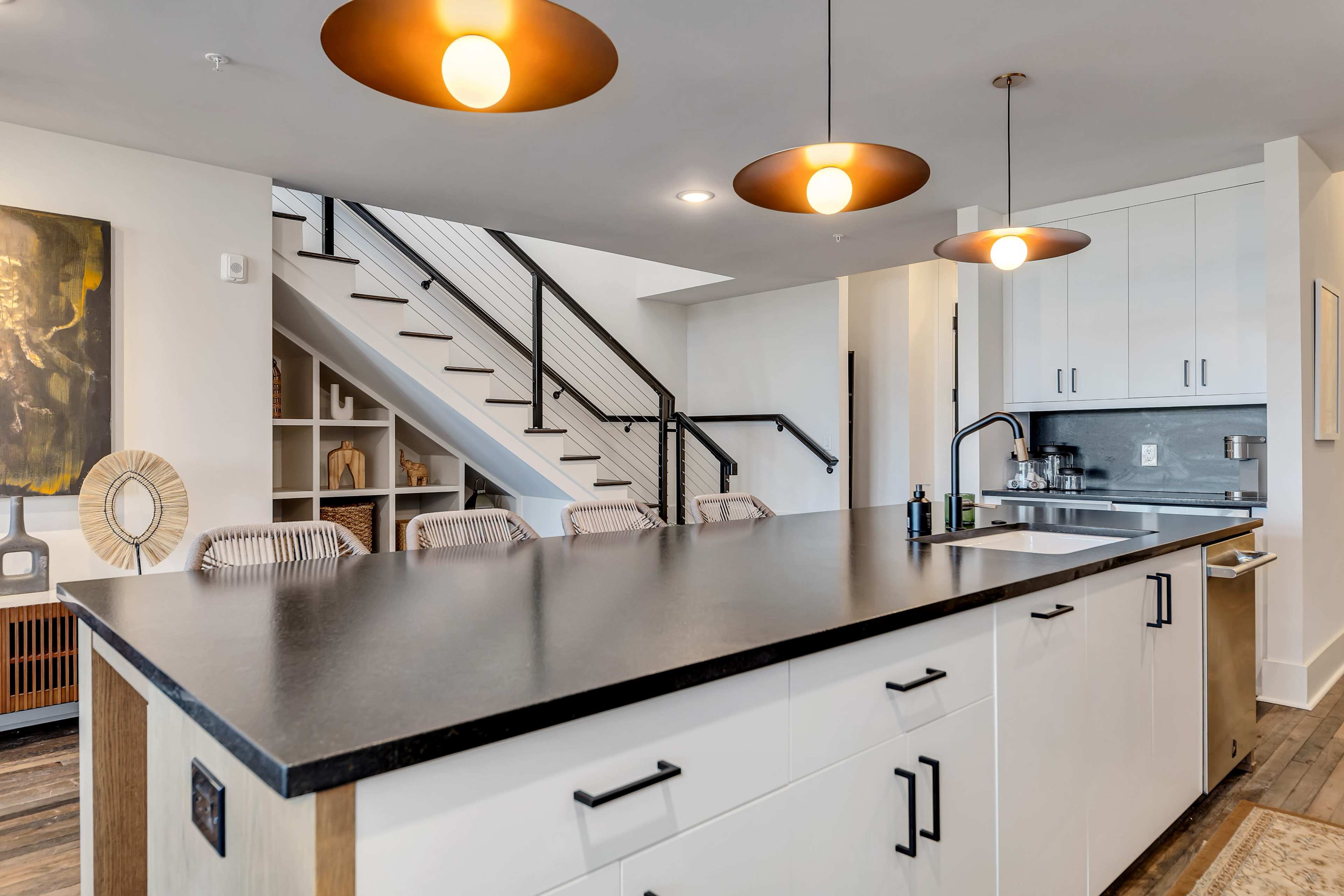 A modern kitchen features a large black countertop, white cabinets, and pendant lights above seating near a staircase.