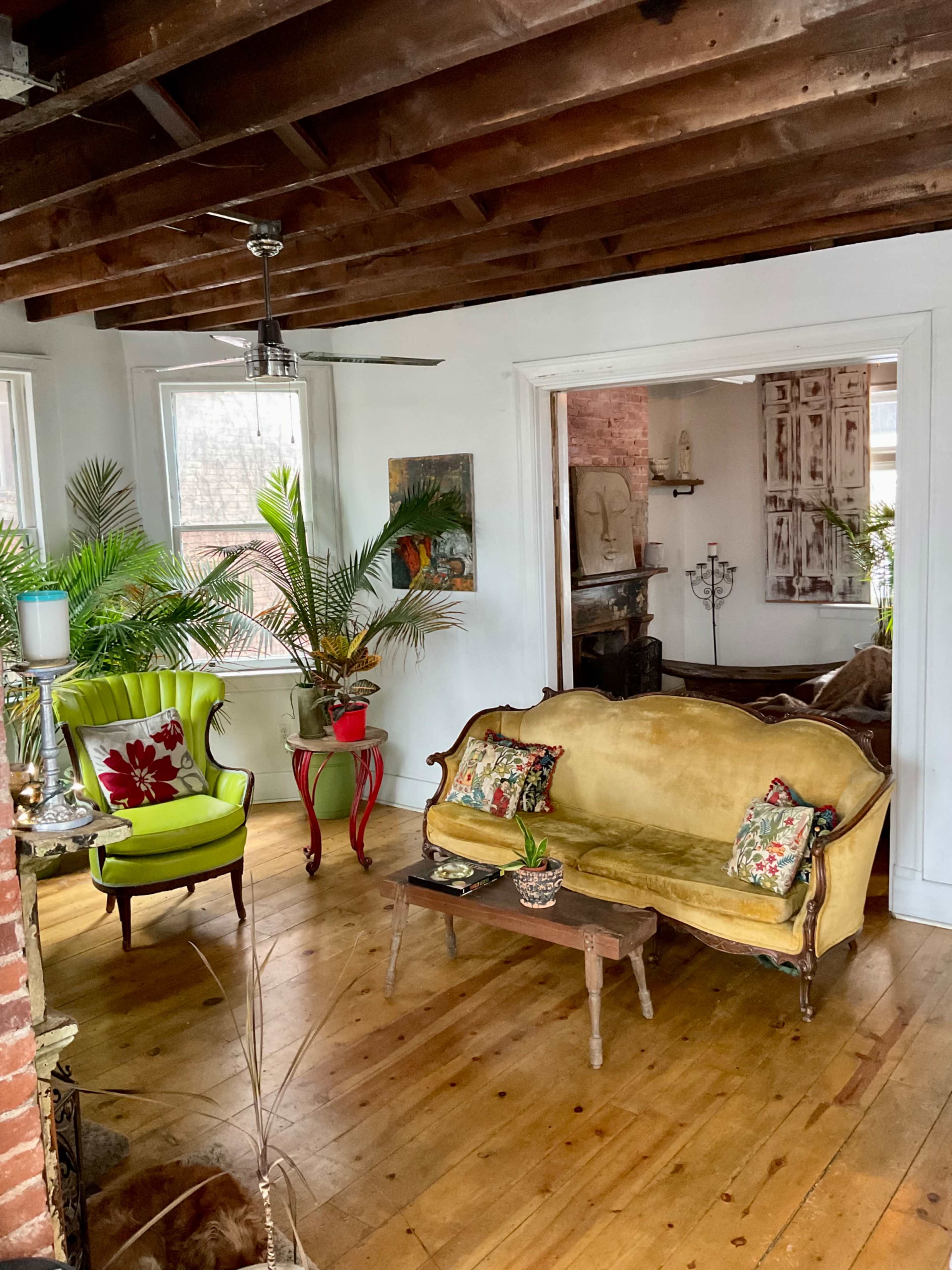 A cozy living room with a yellow sofa, a green armchair, and decorative plants, set against exposed wooden beams and brick walls.