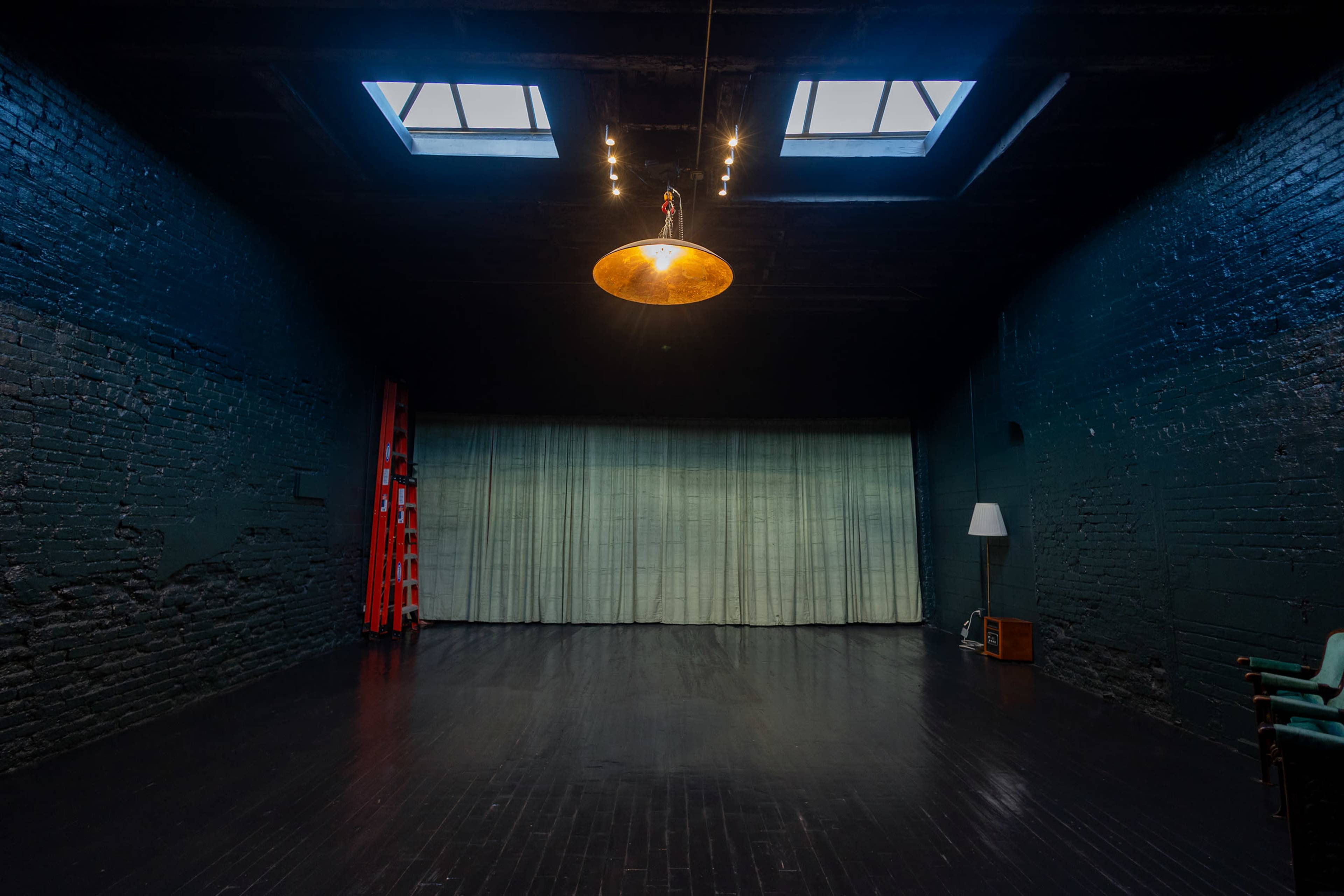 A spacious, dimly lit room with dark walls, a wooden floor, a suspended light fixture, a green curtain, a ladder, and a lamp in the corner.
