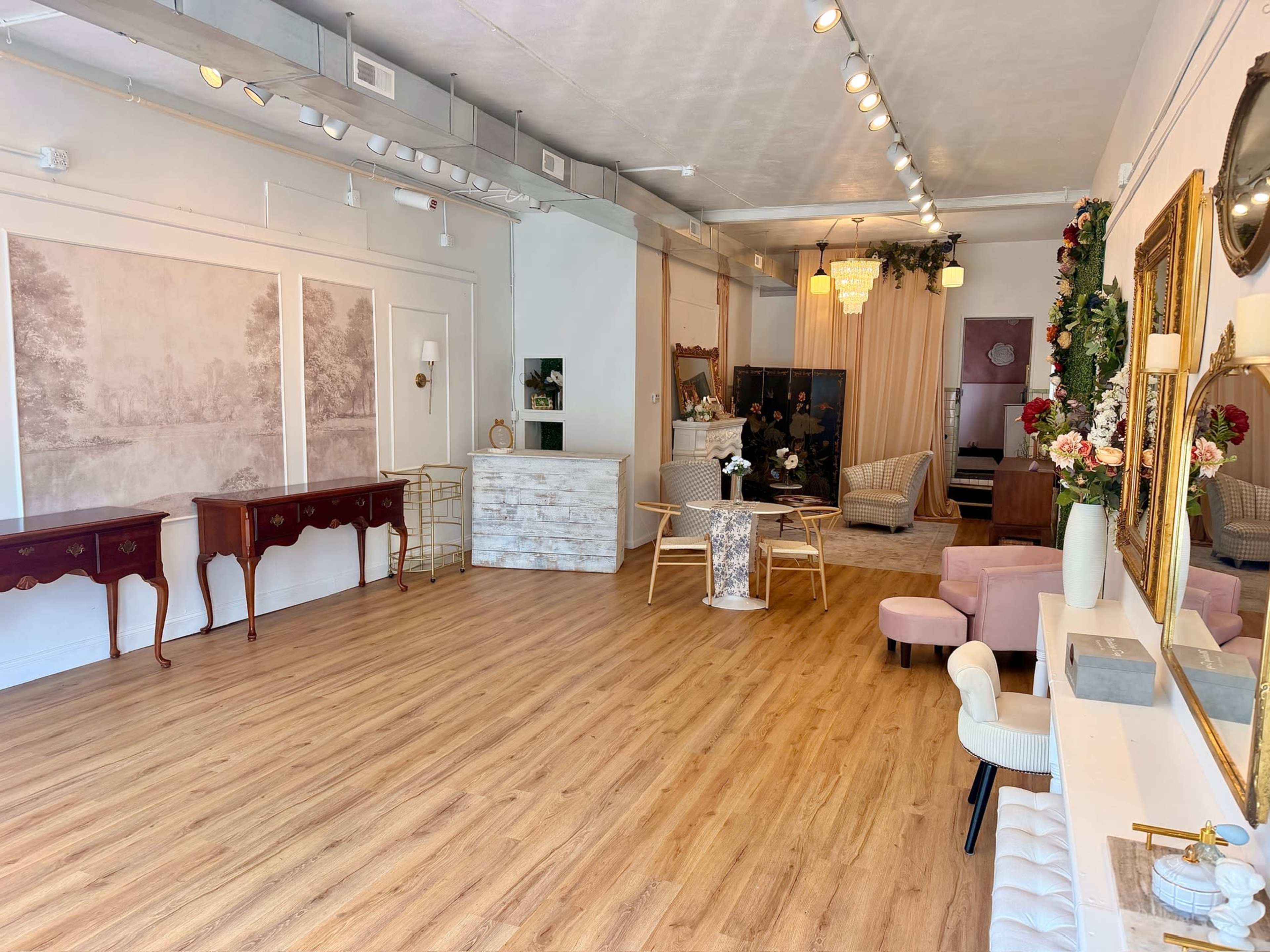 The image shows a spacious, well-lit interior featuring wooden flooring, elegant furnishings, and decorative elements such as a mirror and floral arrangements.