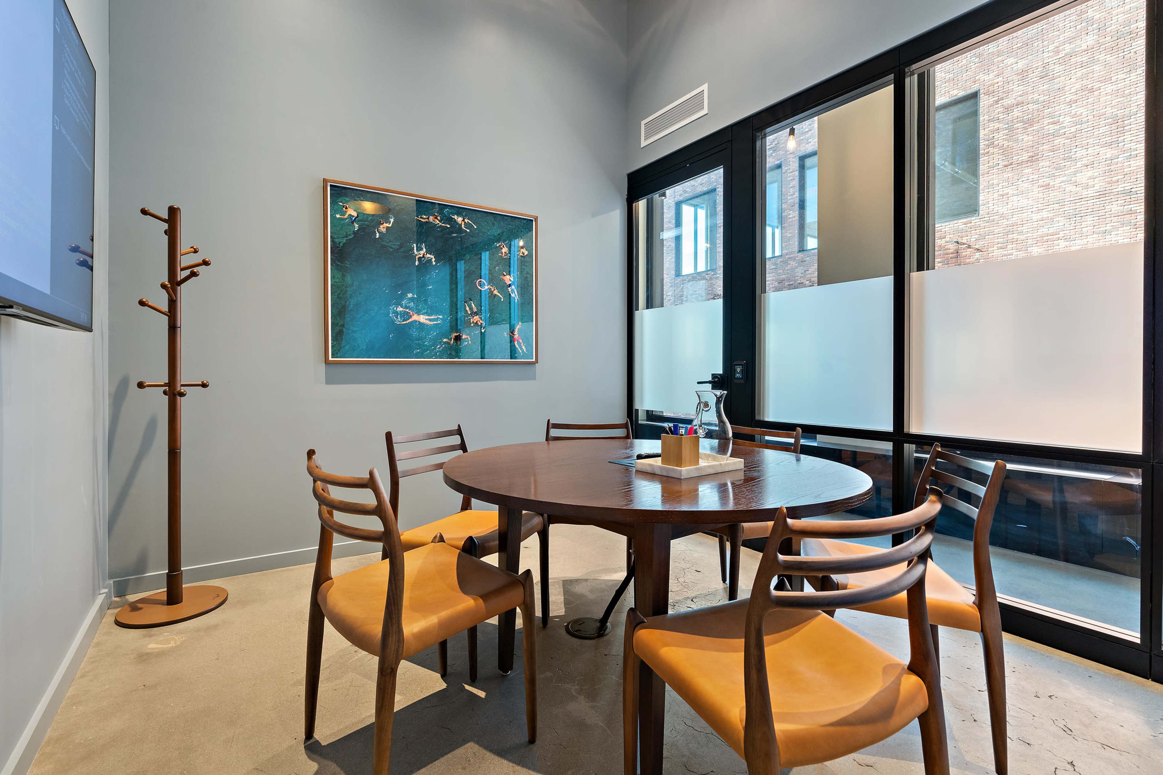 A small conference room features a round wooden table surrounded by four wooden chairs, a coat rack, and a large artwork on the wall.