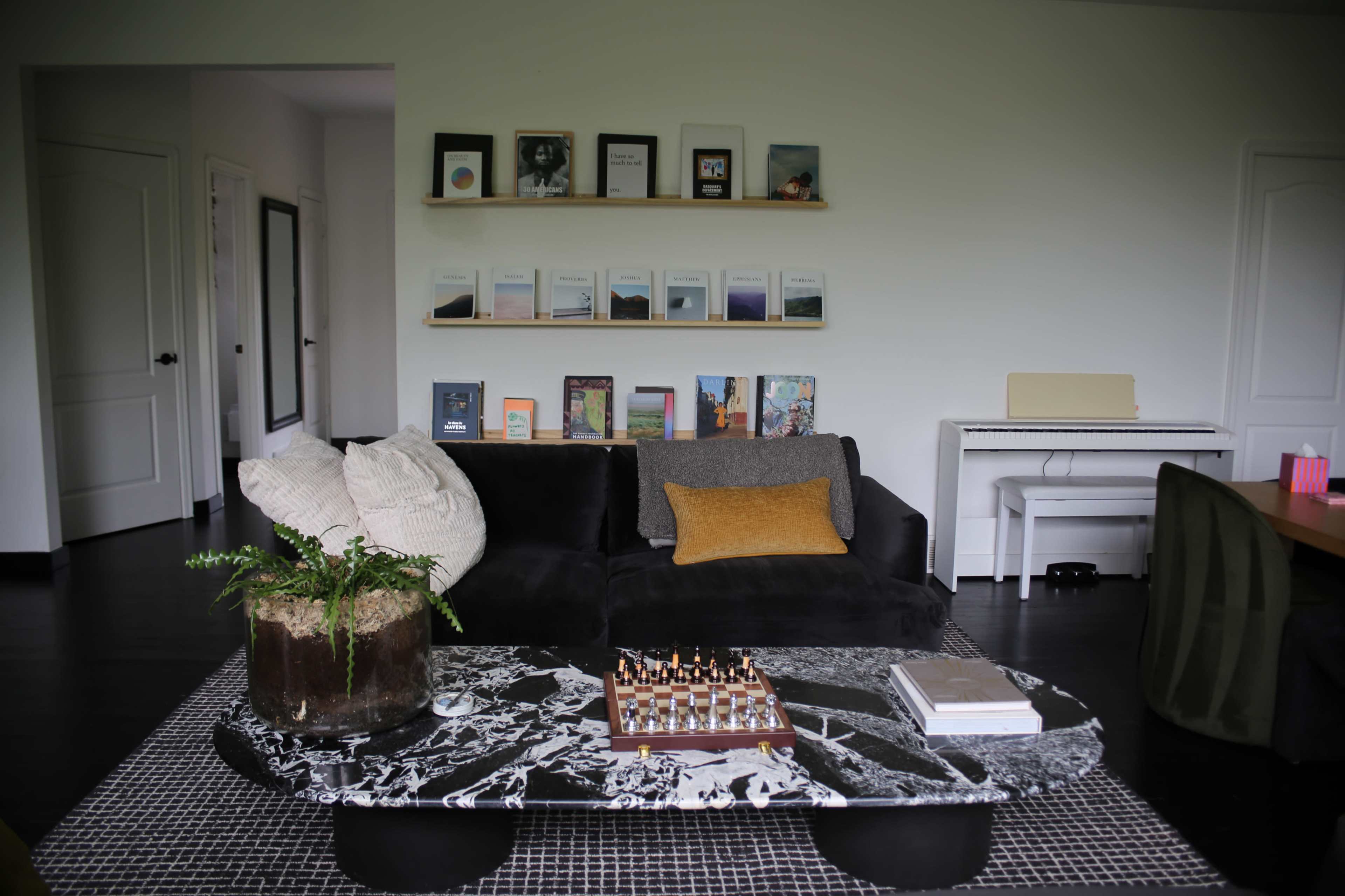 A modern living room features a black sofa with decorative pillows, a marble coffee table with a chess set, and a gallery wall of framed artworks and books.