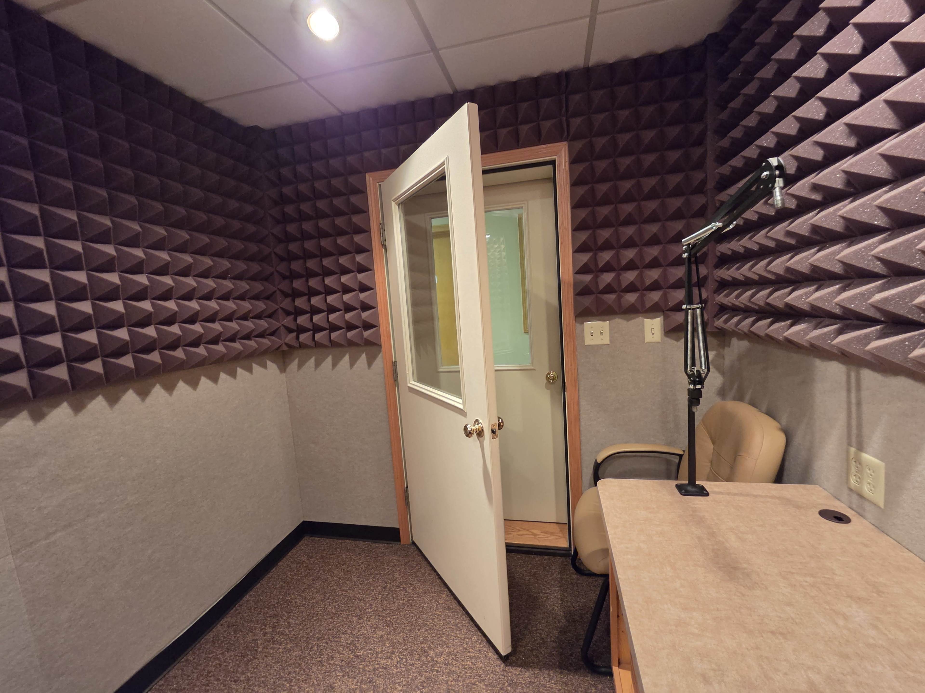 The image shows a soundproof recording studio with purple acoustic foam on the walls, a beige office chair, a small table, and a partially open door.