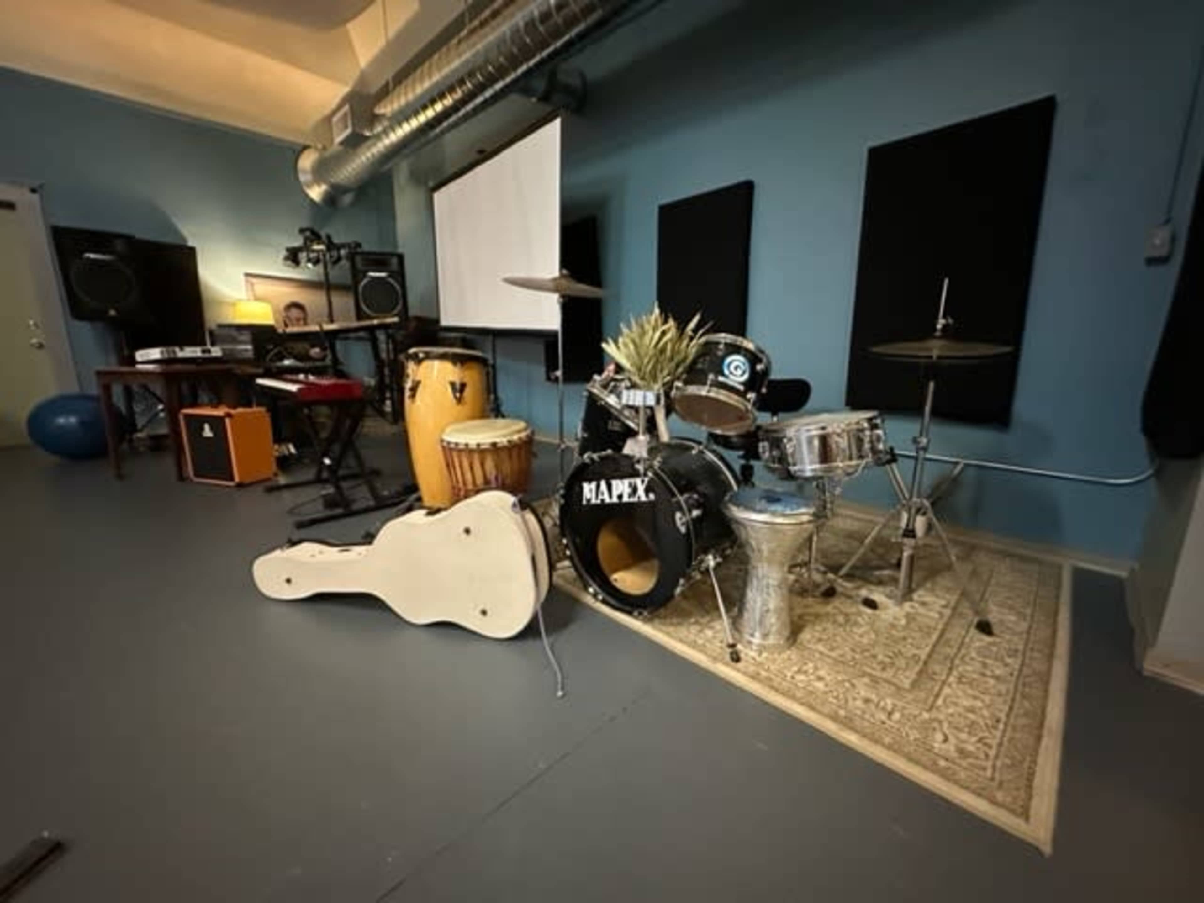 The image shows a music studio with various instruments including a drum set, congas, a guitar, and audio equipment against blue walls.