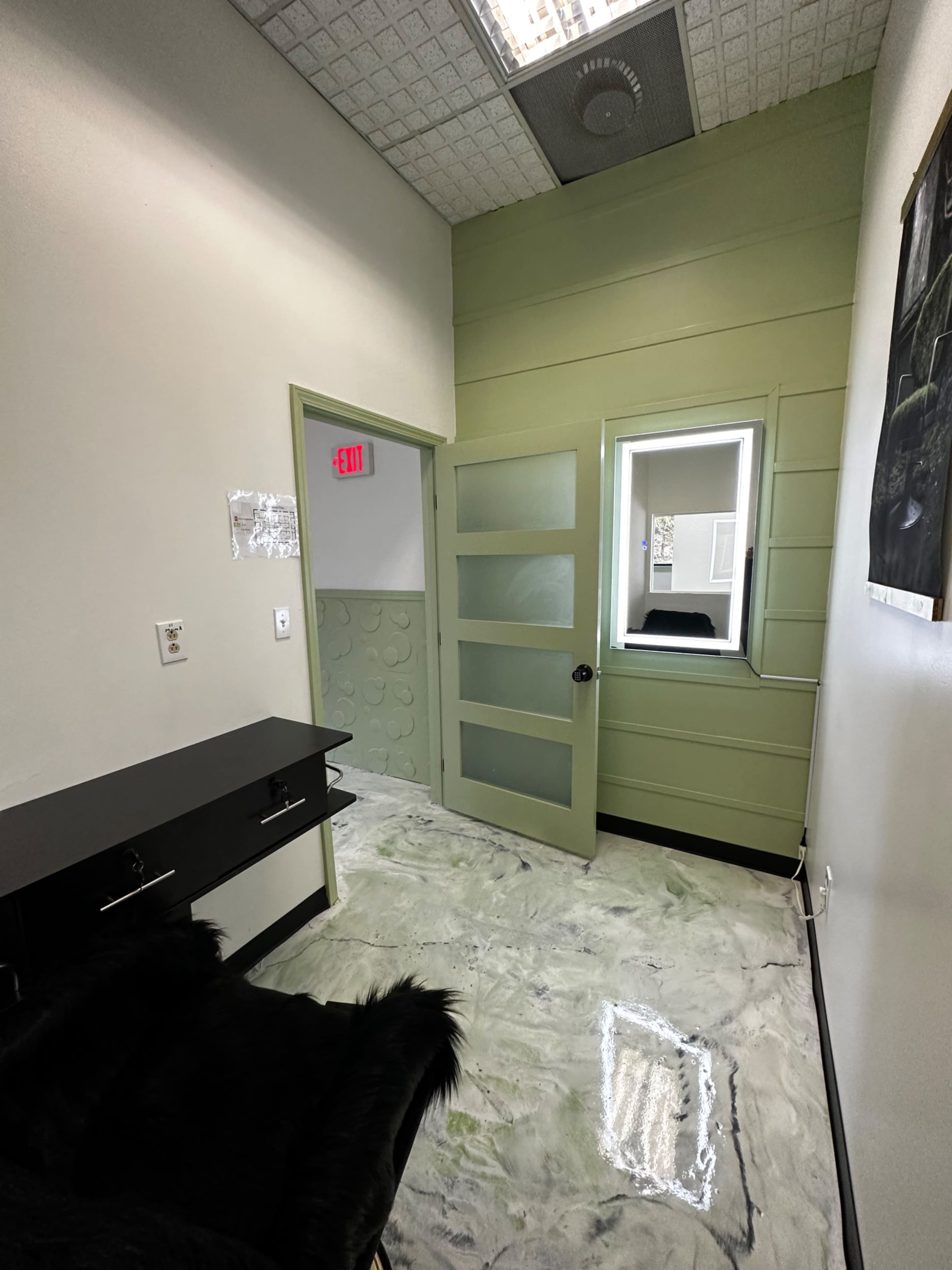 The image shows a small, well-lit room with green walls, a desk, and a door leading to another space.