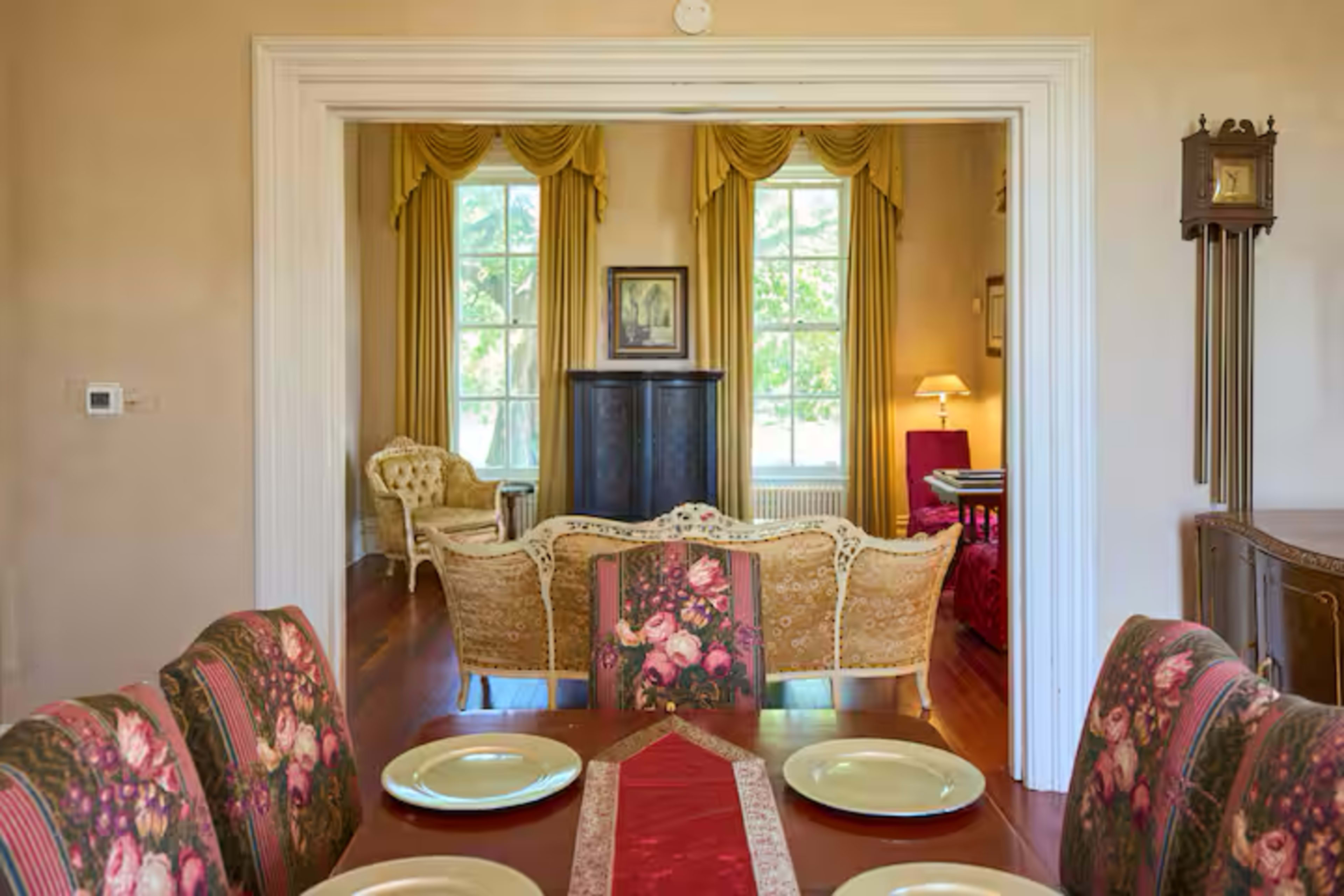 A dining room with a floral table setting is framed by an archway leading to a living area that features large windows and elegant furniture.