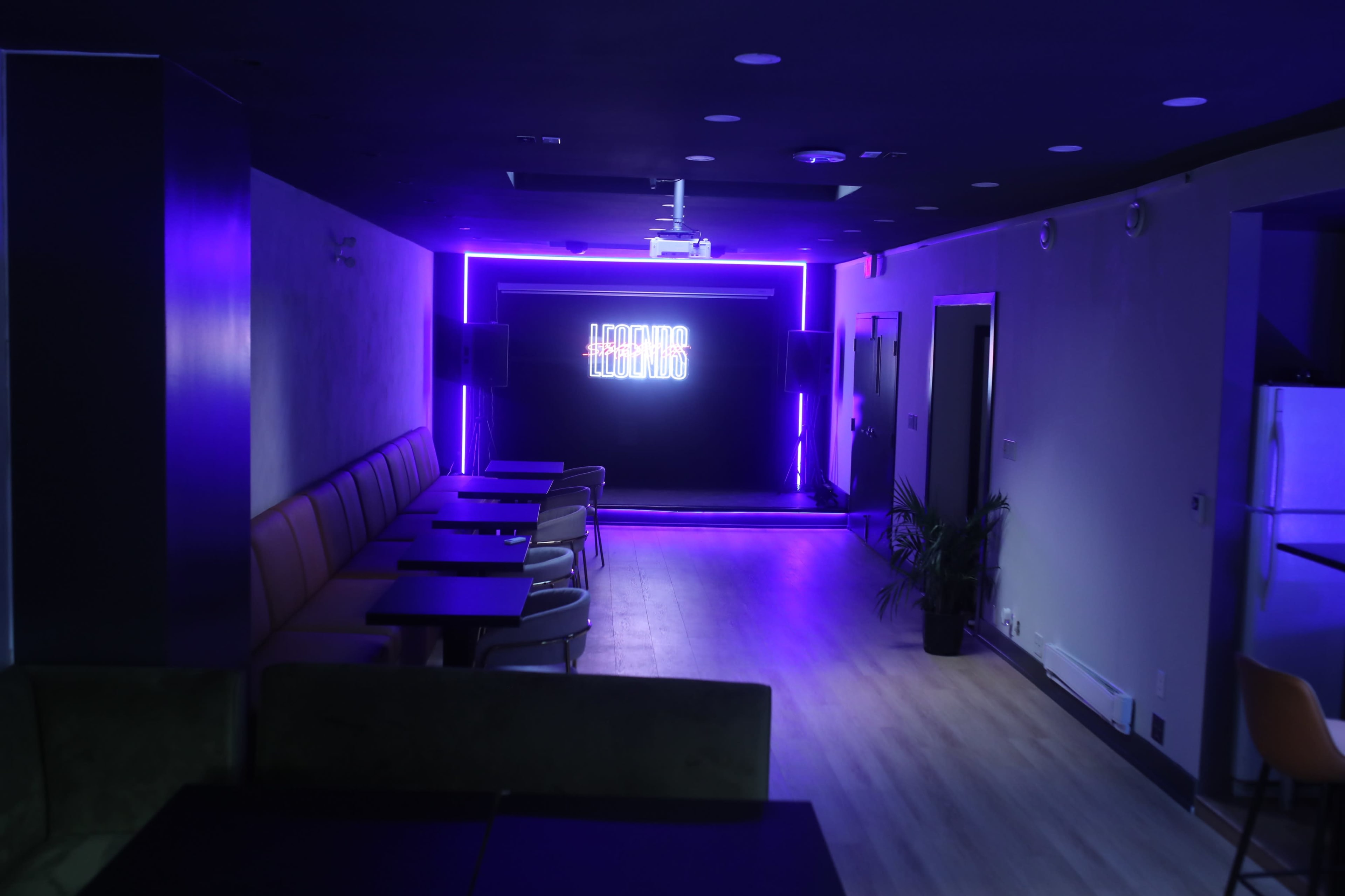 The image shows a dimly lit lounge with purple neon lighting, featuring wooden flooring, tables, and chairs arranged in an open layout.