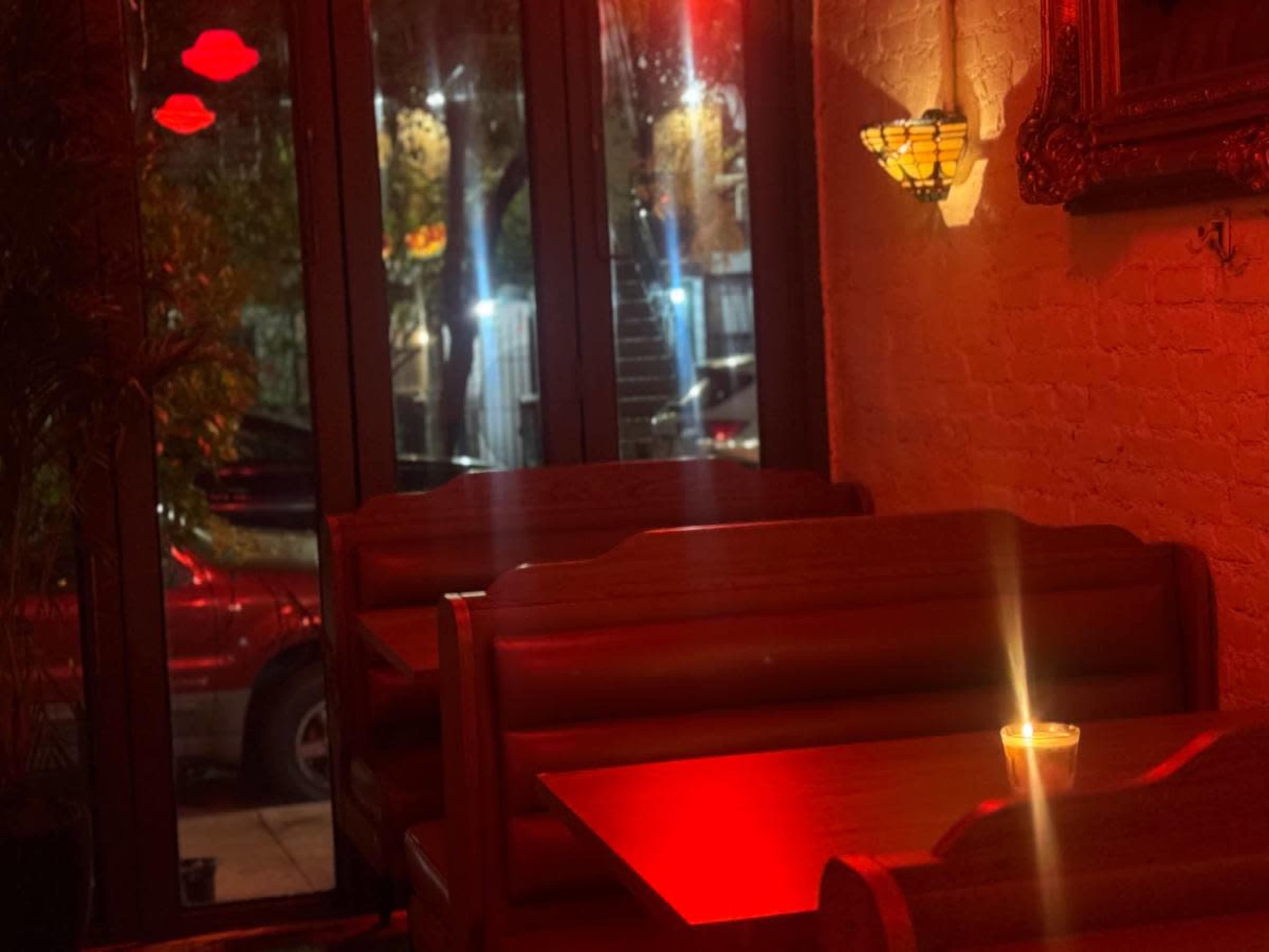 A dimly lit restaurant interior features wooden booths and a candle on a table, with red lighting and large windows showing the street outside.