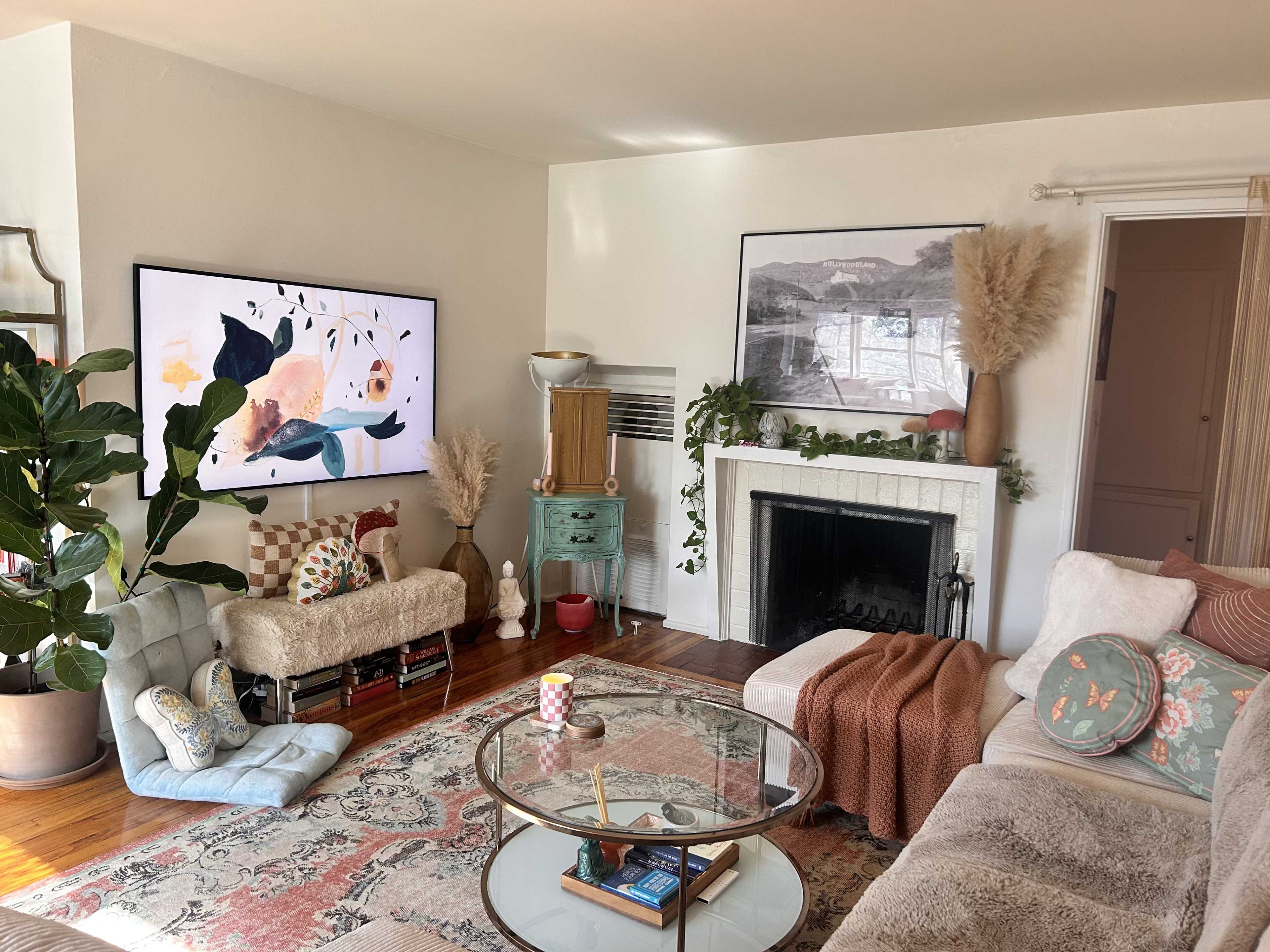 A living room features a large television mounted on the wall displaying abstract art, a green accent table, a fireplace, and various plants and decorative cushions.