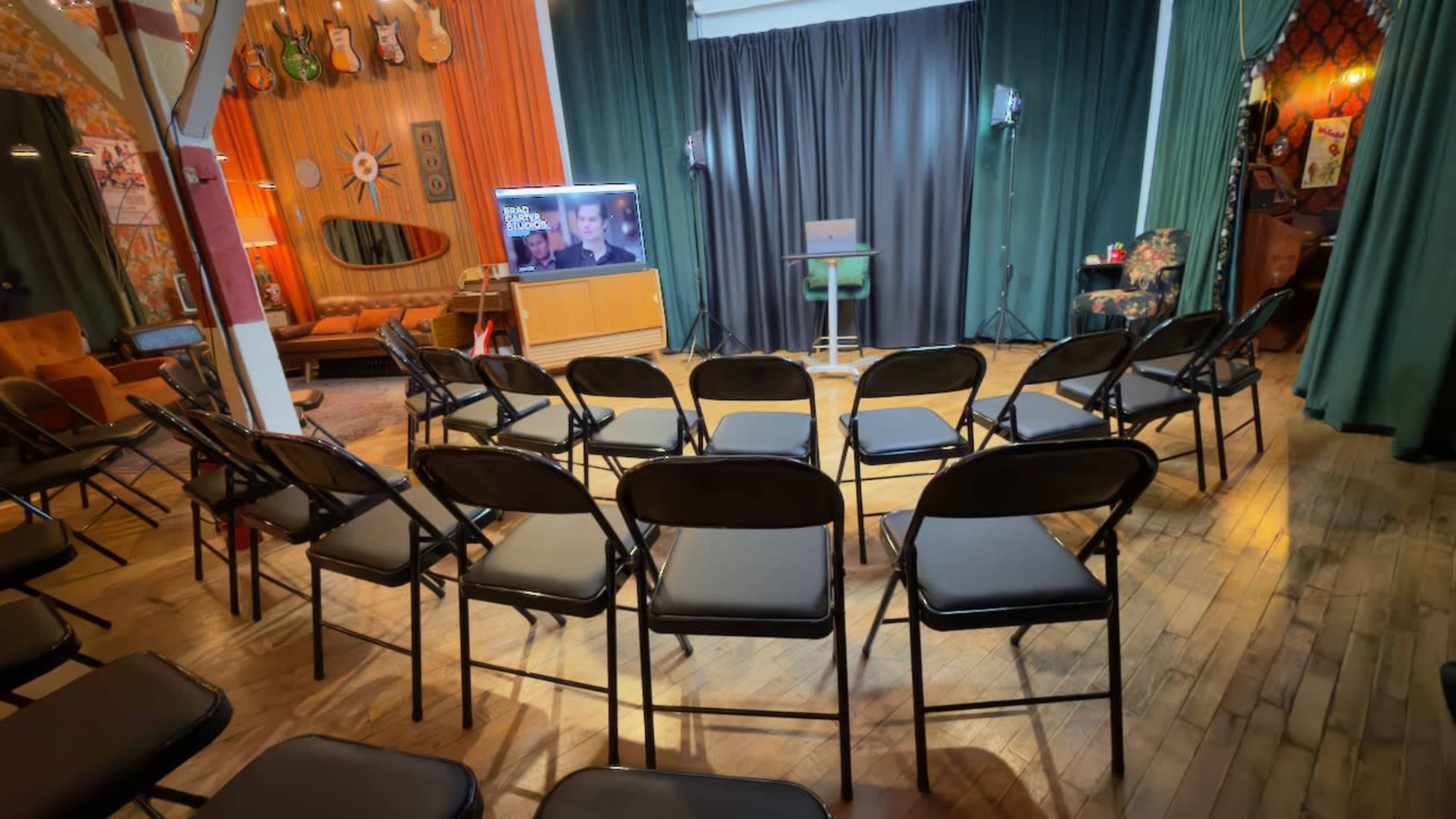 The image shows a room set up for a presentation, featuring rows of black chairs facing a small stage with a monitor and a green stool.