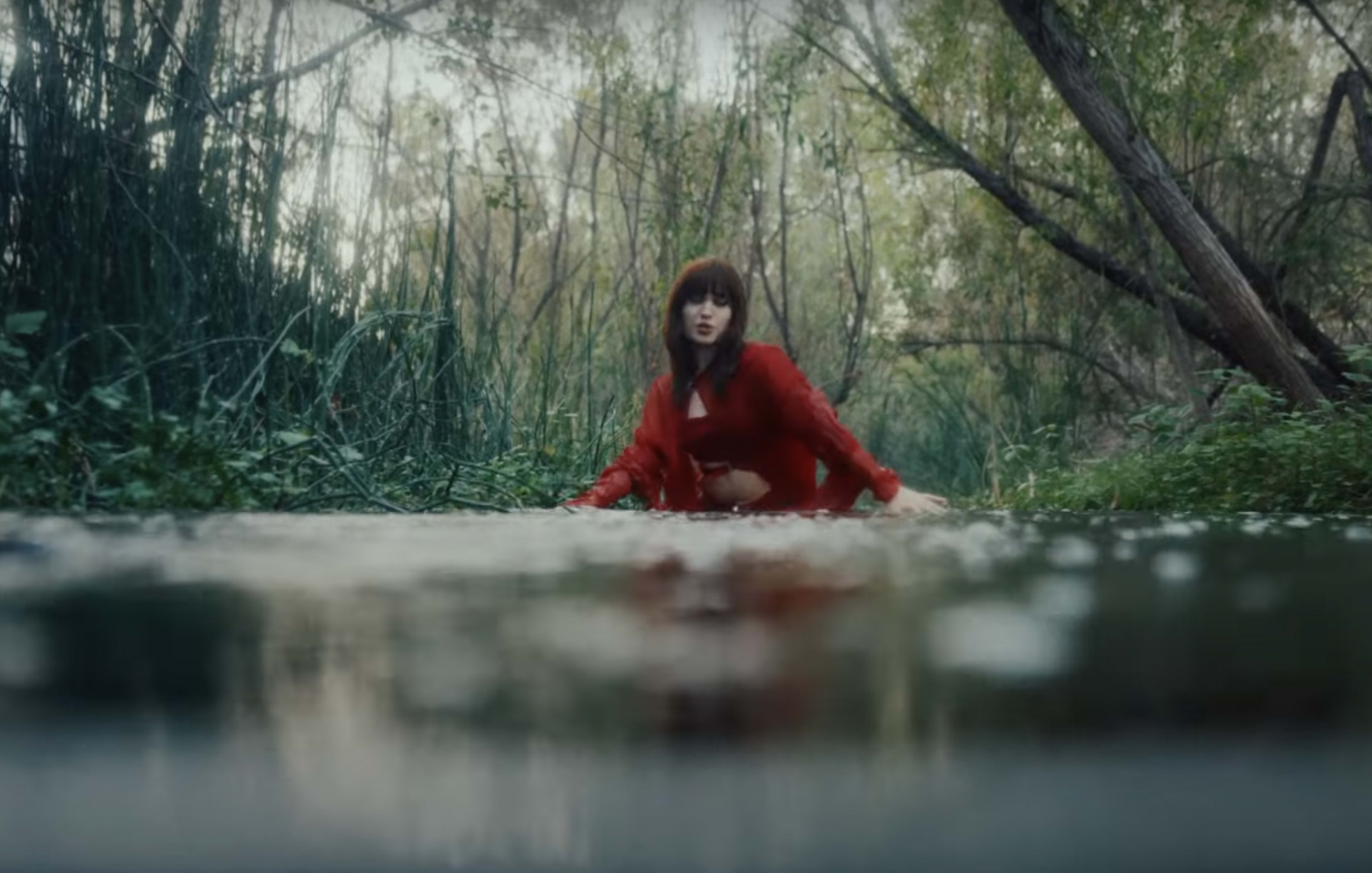 A woman in a red outfit sits in shallow water surrounded by dense greenery and trees.