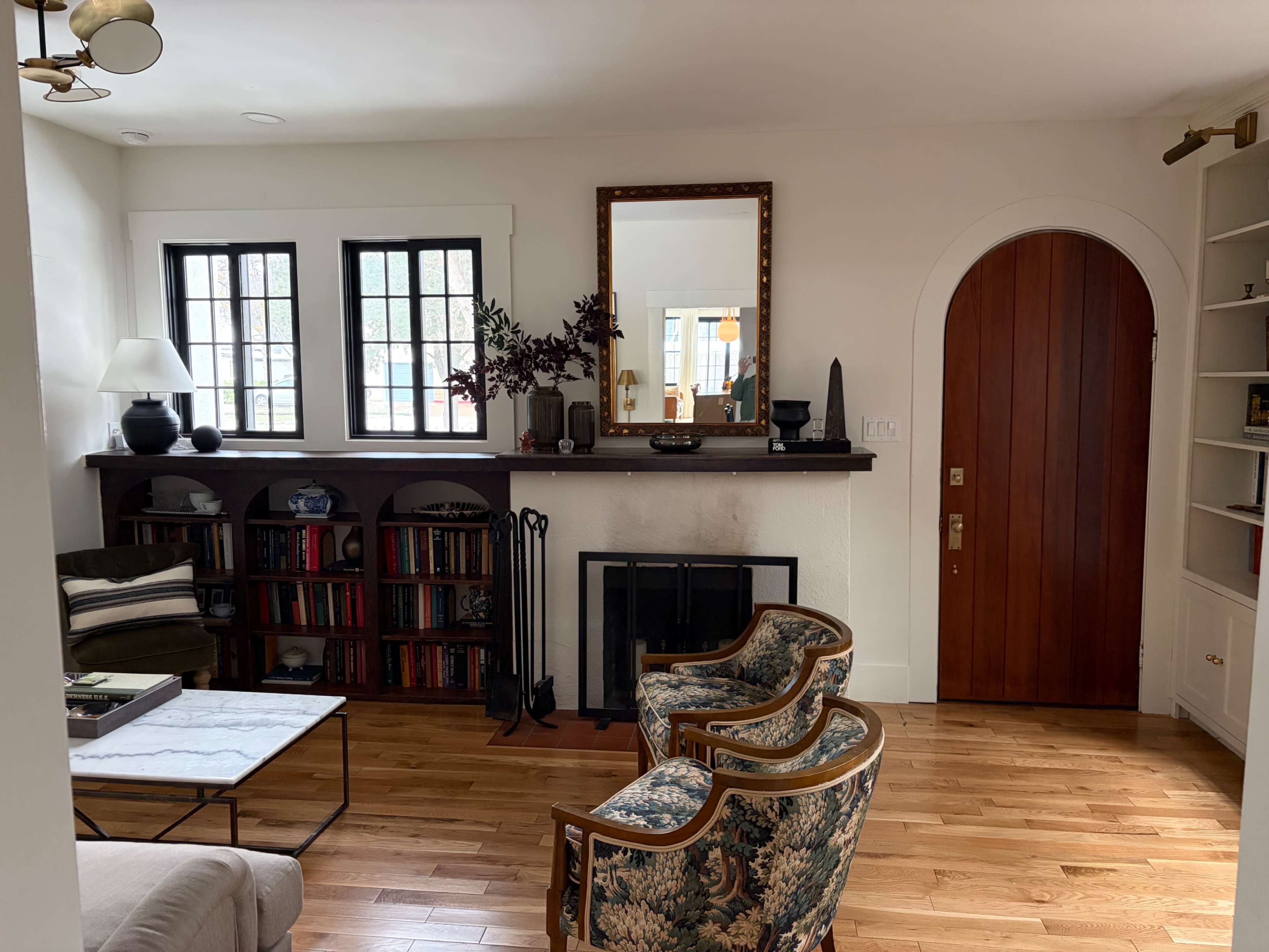 The image shows a living room with wooden floors, a bookshelf filled with books, two patterned chairs facing a coffee table, and a fireplace against a white wall.