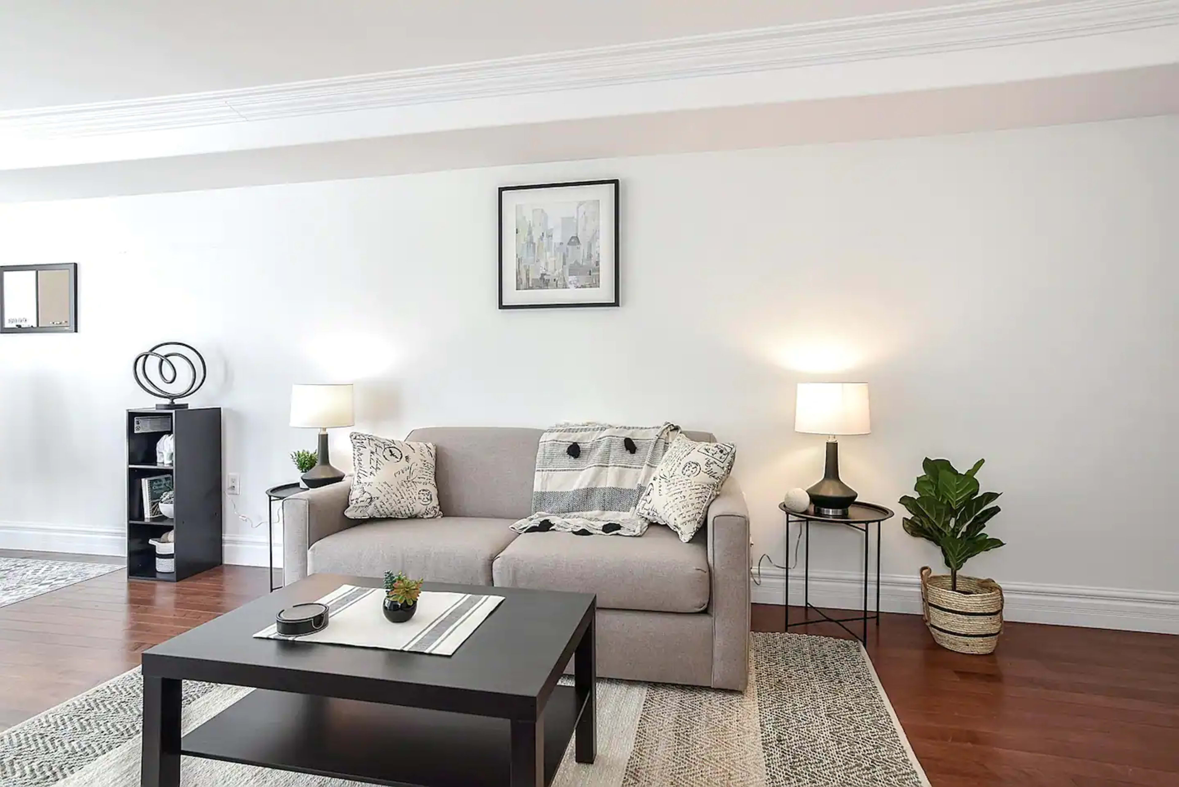 A modern living room with a gray sofa, a black coffee table, and two lamps on either side, featuring a plant in the corner and a framed cityscape on the wall.
