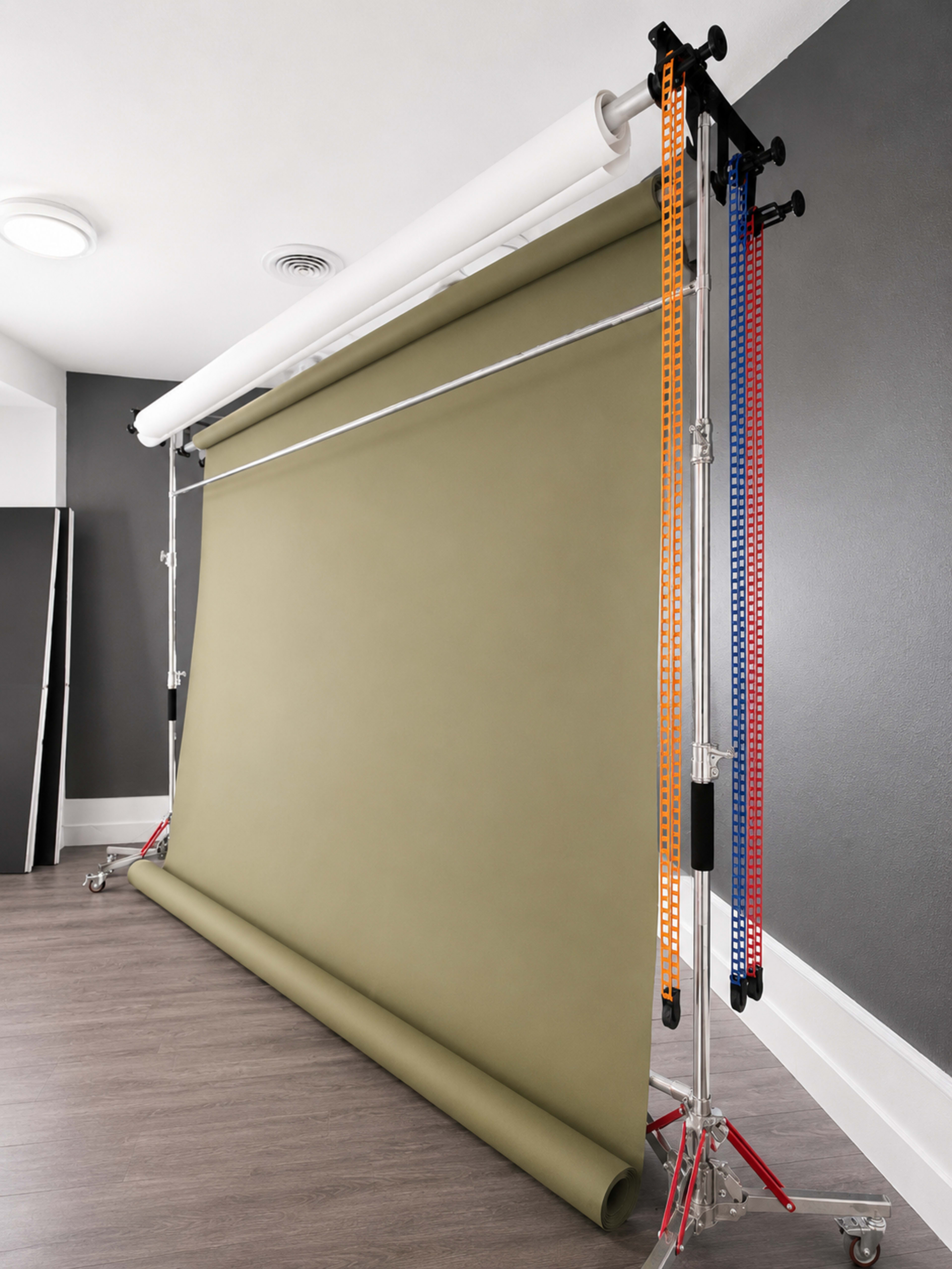The image shows a photography backdrop setup, featuring a rolled olive-green backdrop on a support stand in a room with gray walls and wooden flooring.