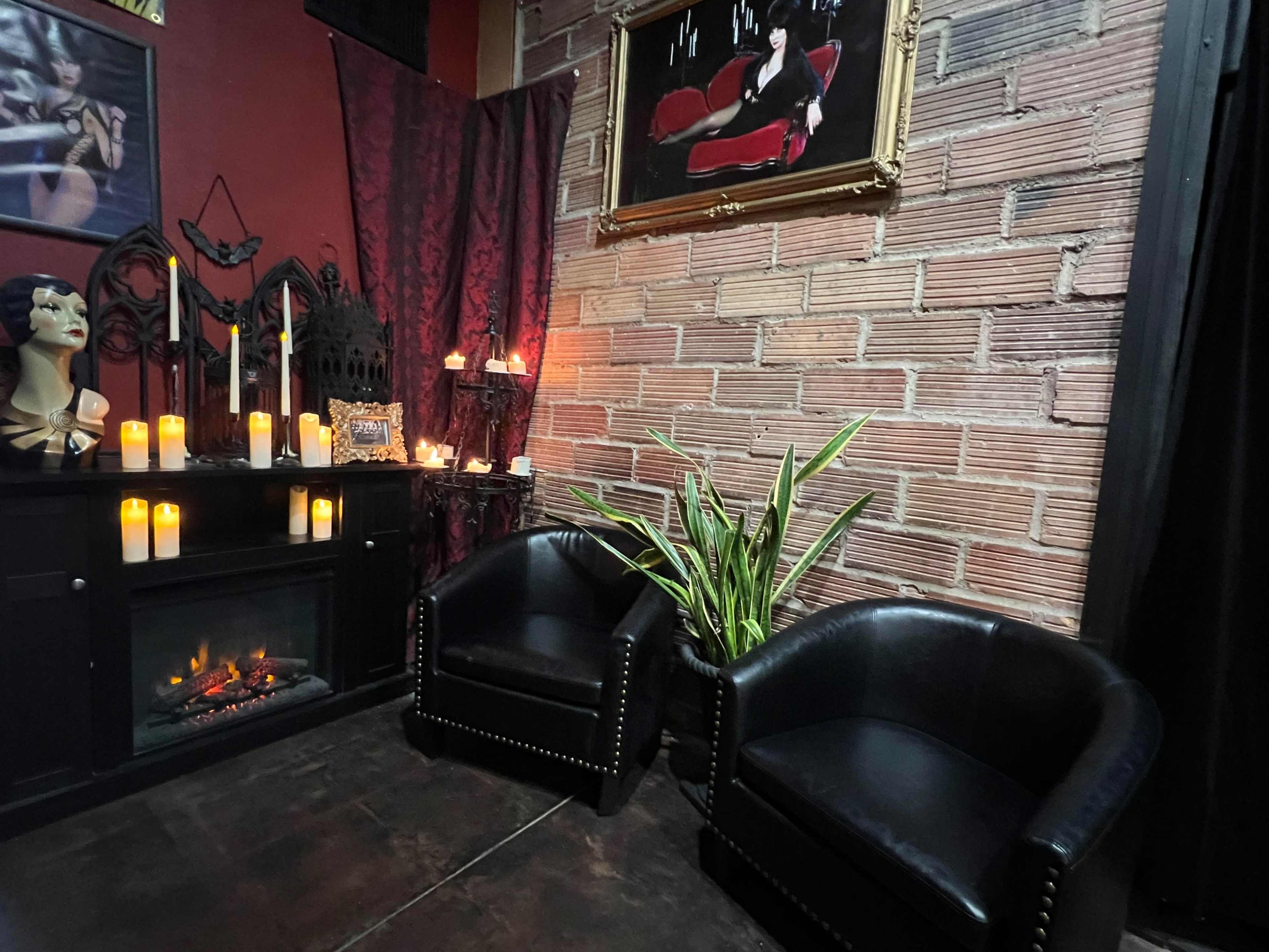 A corner of a dimly lit room features two black leather chairs, a faux fireplace surrounded by candles, and a large framed artwork on the wall.