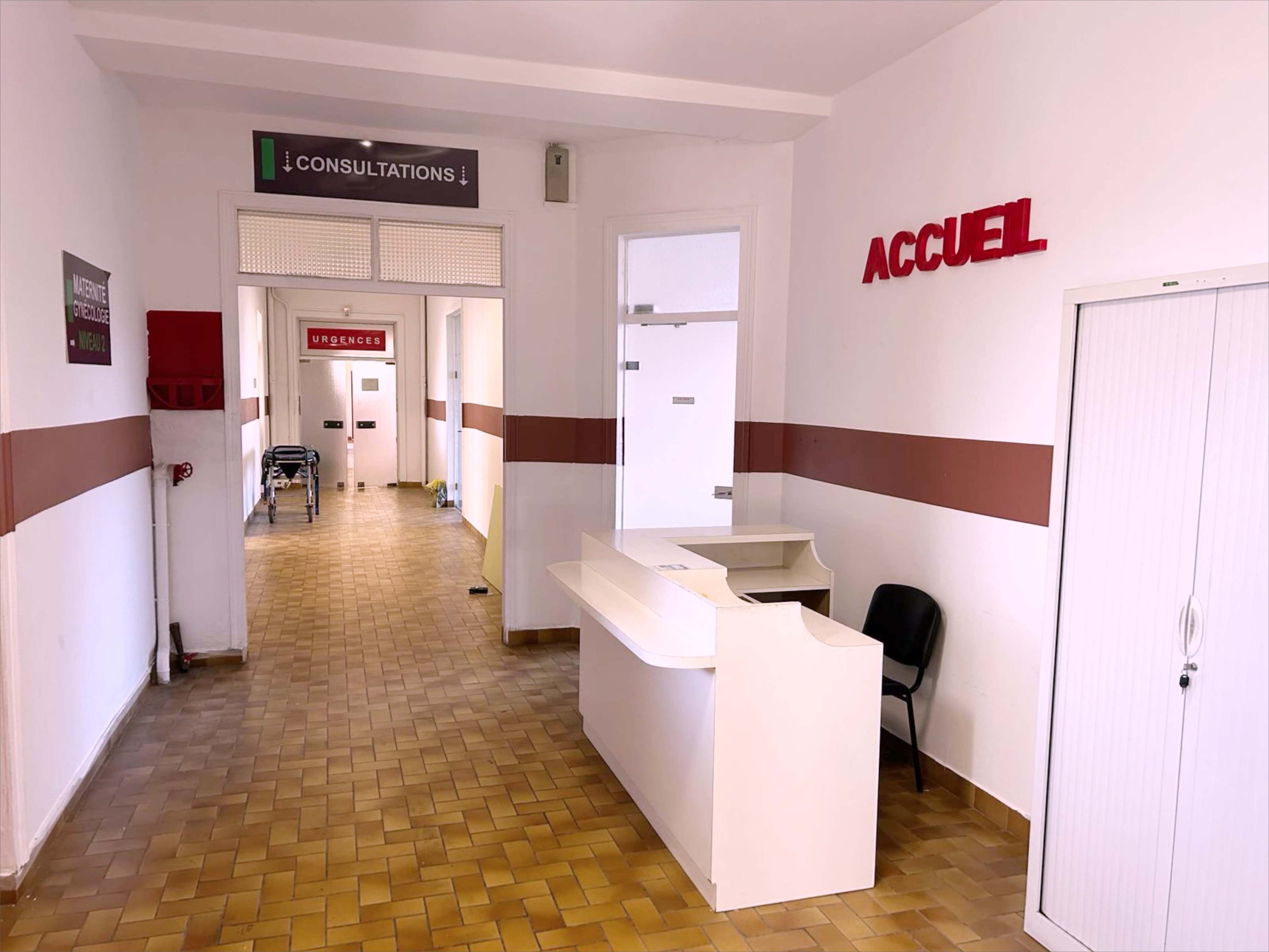 A reception area of a medical facility with a white desk and a black chair, leading to corridors labeled for consultations and emergencies.