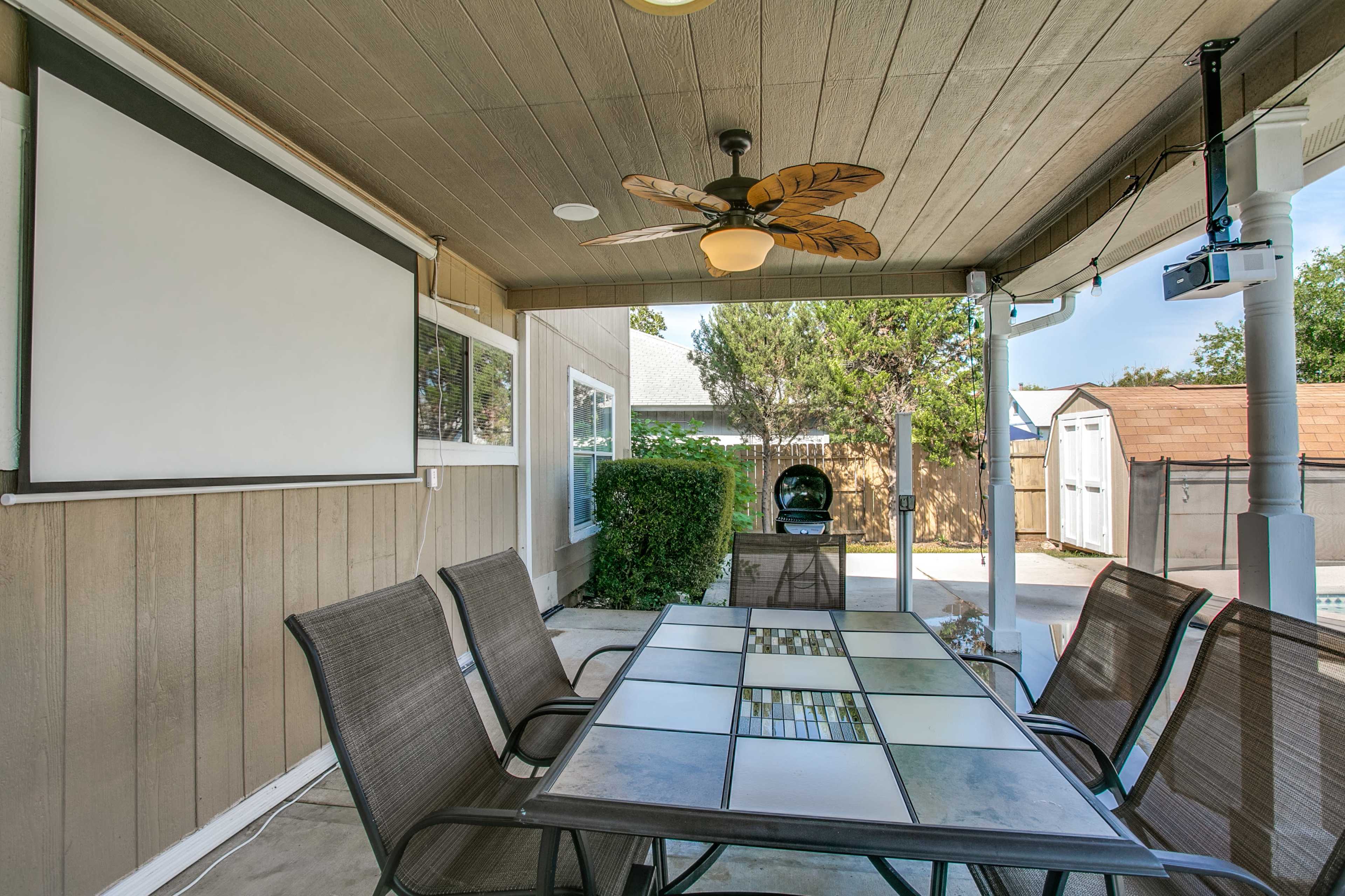 A covered patio area with a dining table, a ceiling fan, and a projector screen, surrounded by greenery and an outdoor shed in the background.
