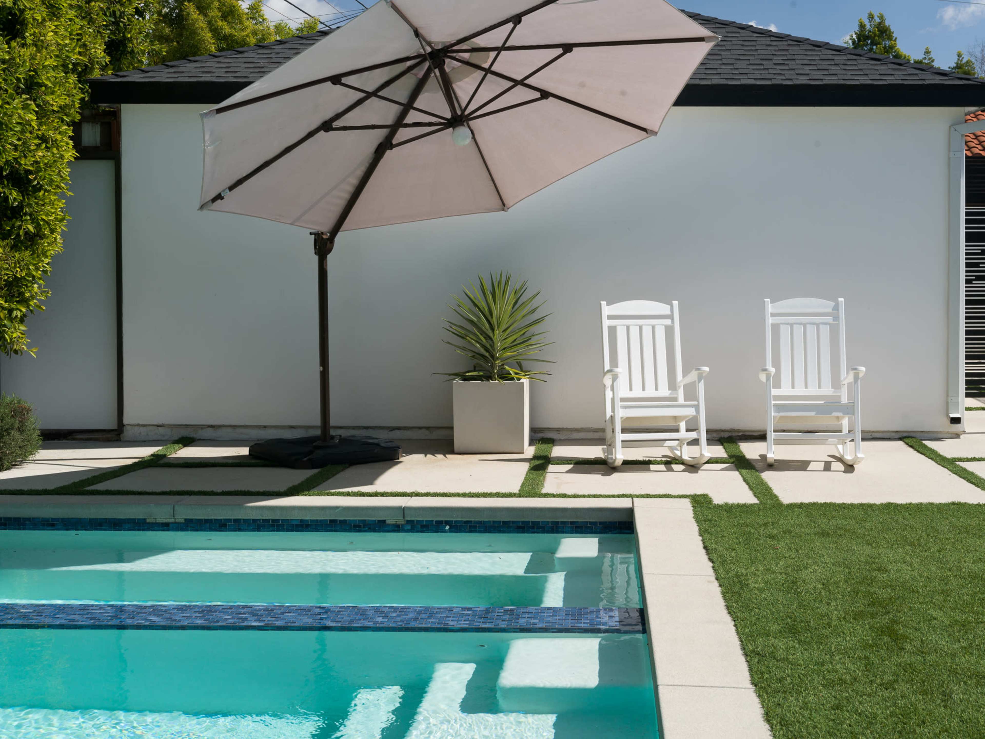 A swimming pool with a light blue water surface is bordered by grass and features a large umbrella and two white rocking chairs nearby.