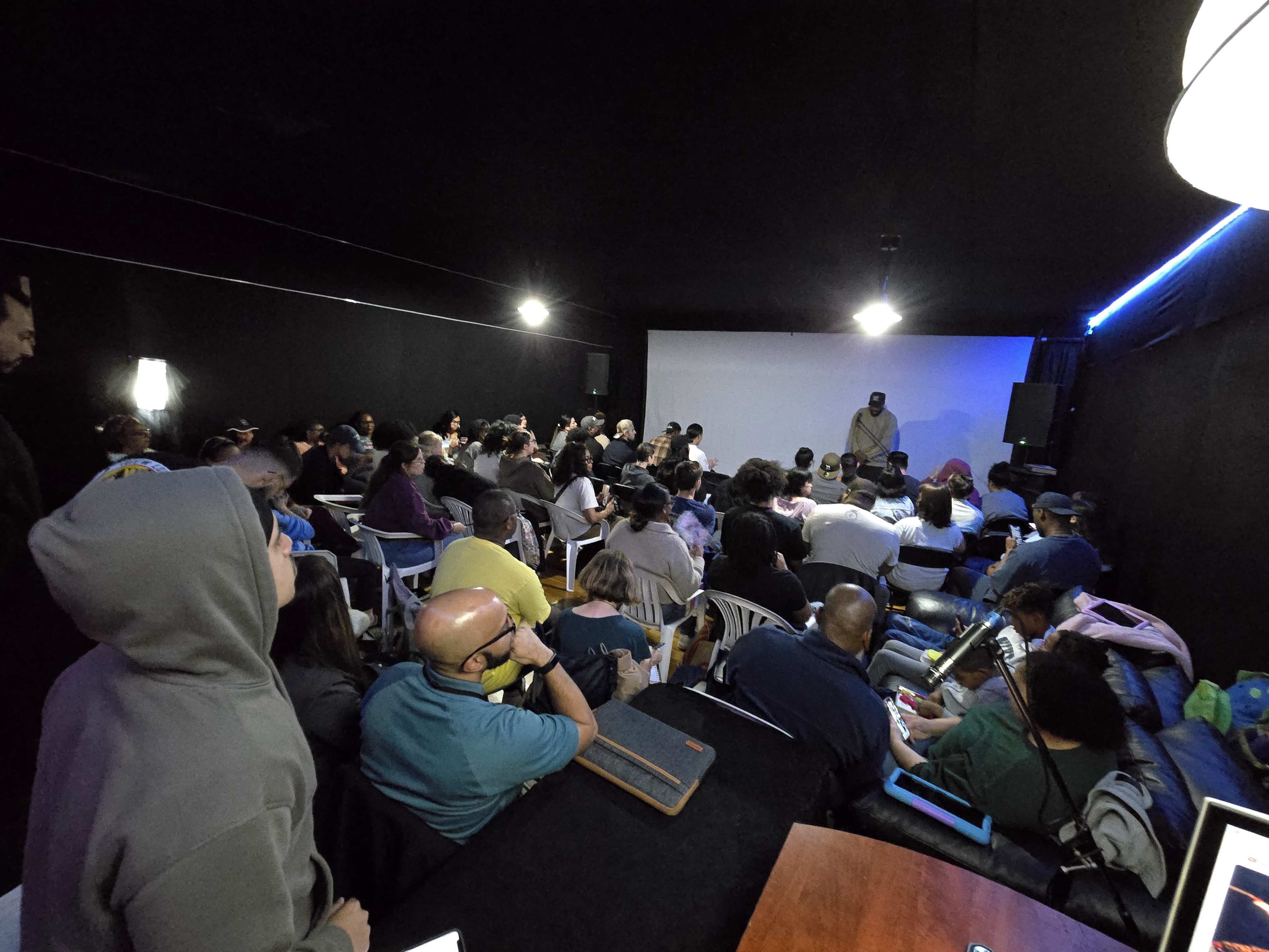 A large audience is seated in a darkened room, facing a speaker in front of a screen.