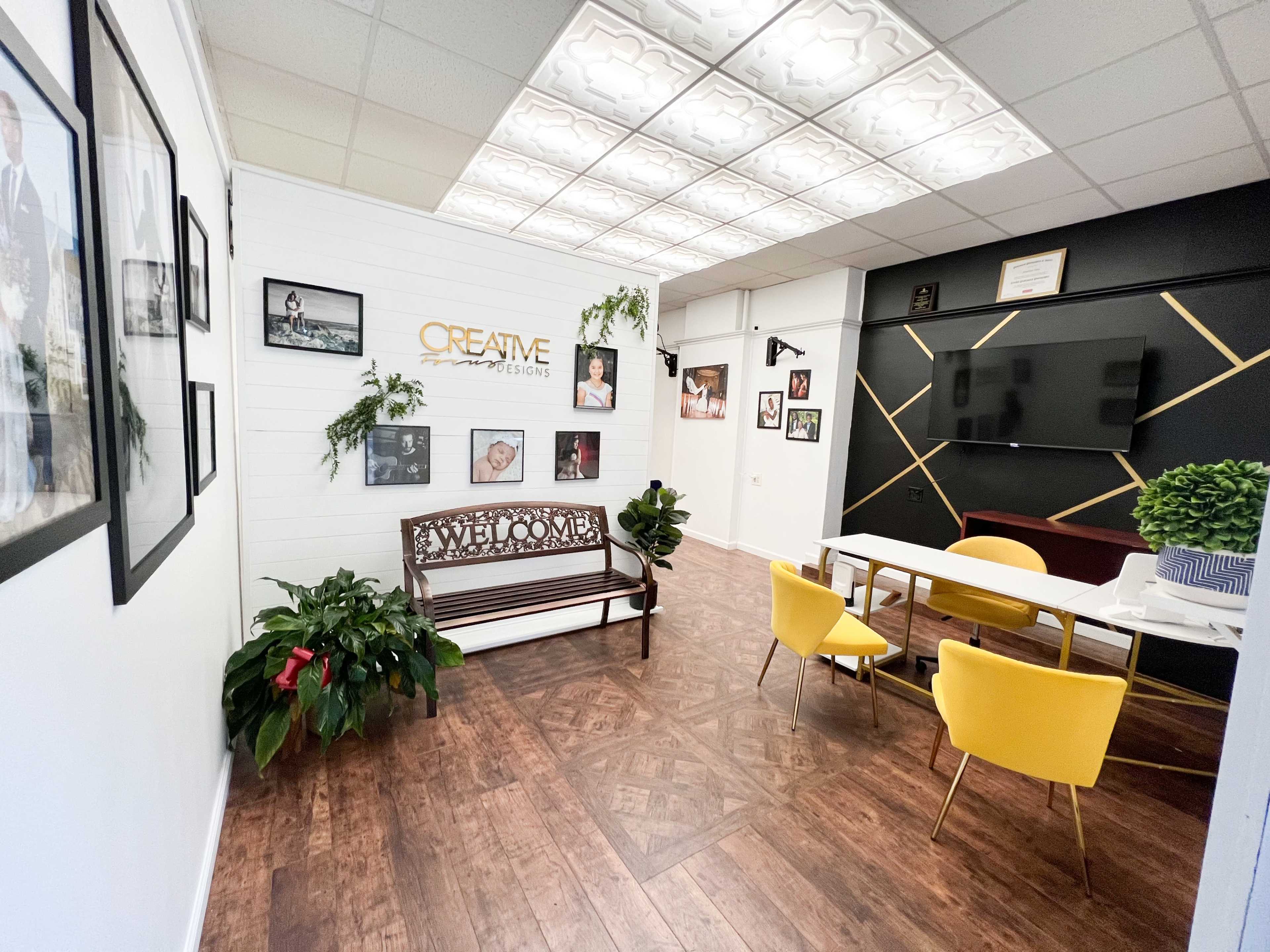 A modern office reception area featuring a wooden bench, yellow chairs, a coffee table, plants, and framed photographs on the walls.
