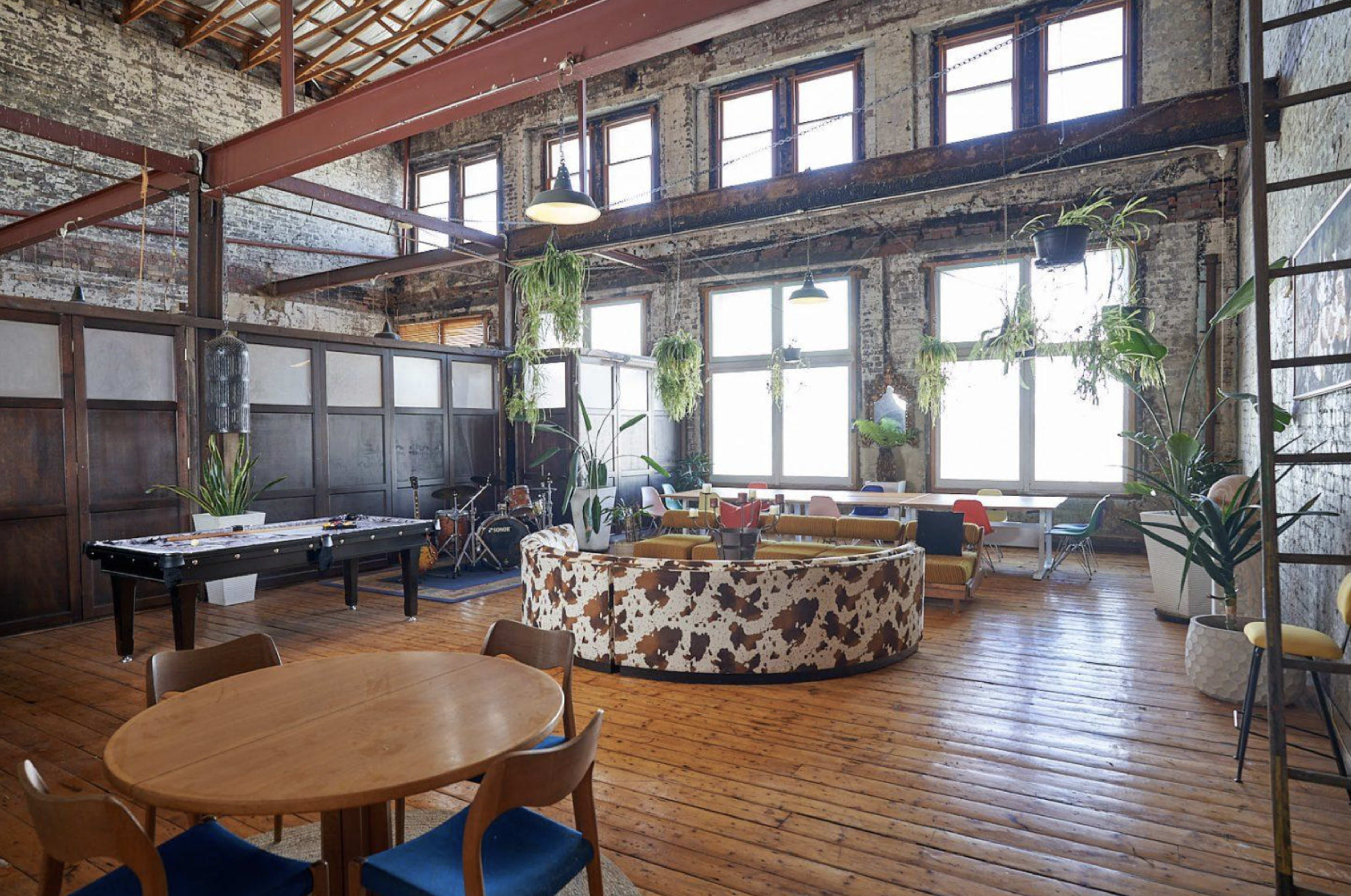 An industrial-style interior with a pool table, a round wooden table, and various seating arrangements surrounded by large windows and hanging plants.
