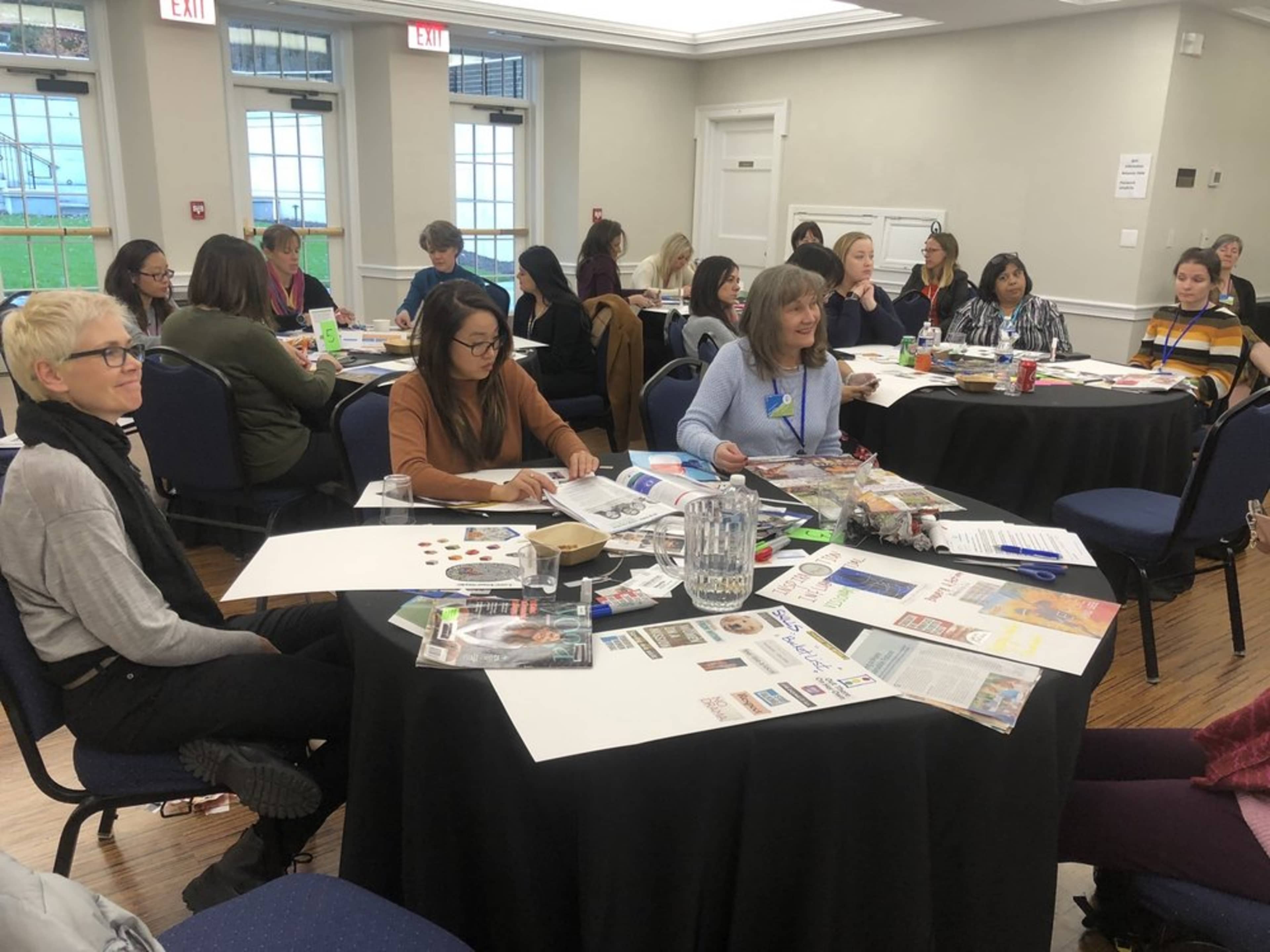 A group of people sits around tables covered with papers, magazines, and craft supplies during a workshop or meeting in a well-lit room.