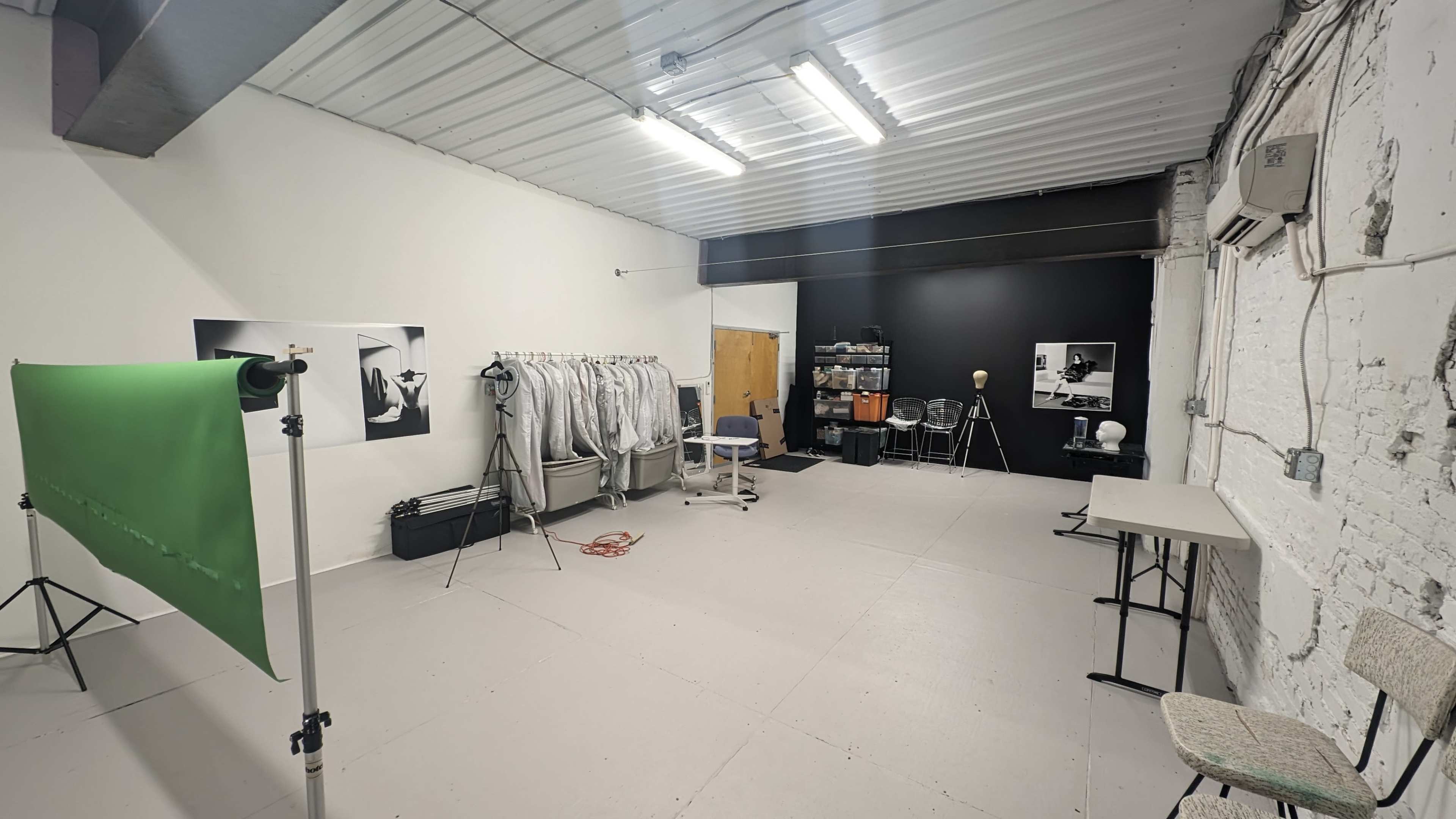 The image shows an empty studio space with white walls, a green backdrop on a stand, and various equipment and furniture along with a black wall at one end.