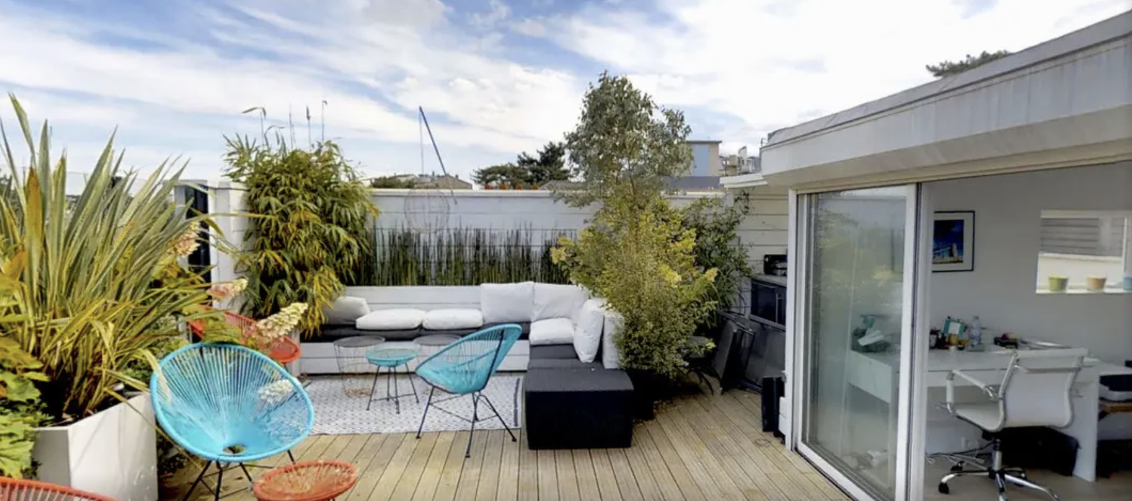 A spacious rooftop terrace features a white sectional sofa, colorful chairs, plants, and a glass door leading into an interior space.
