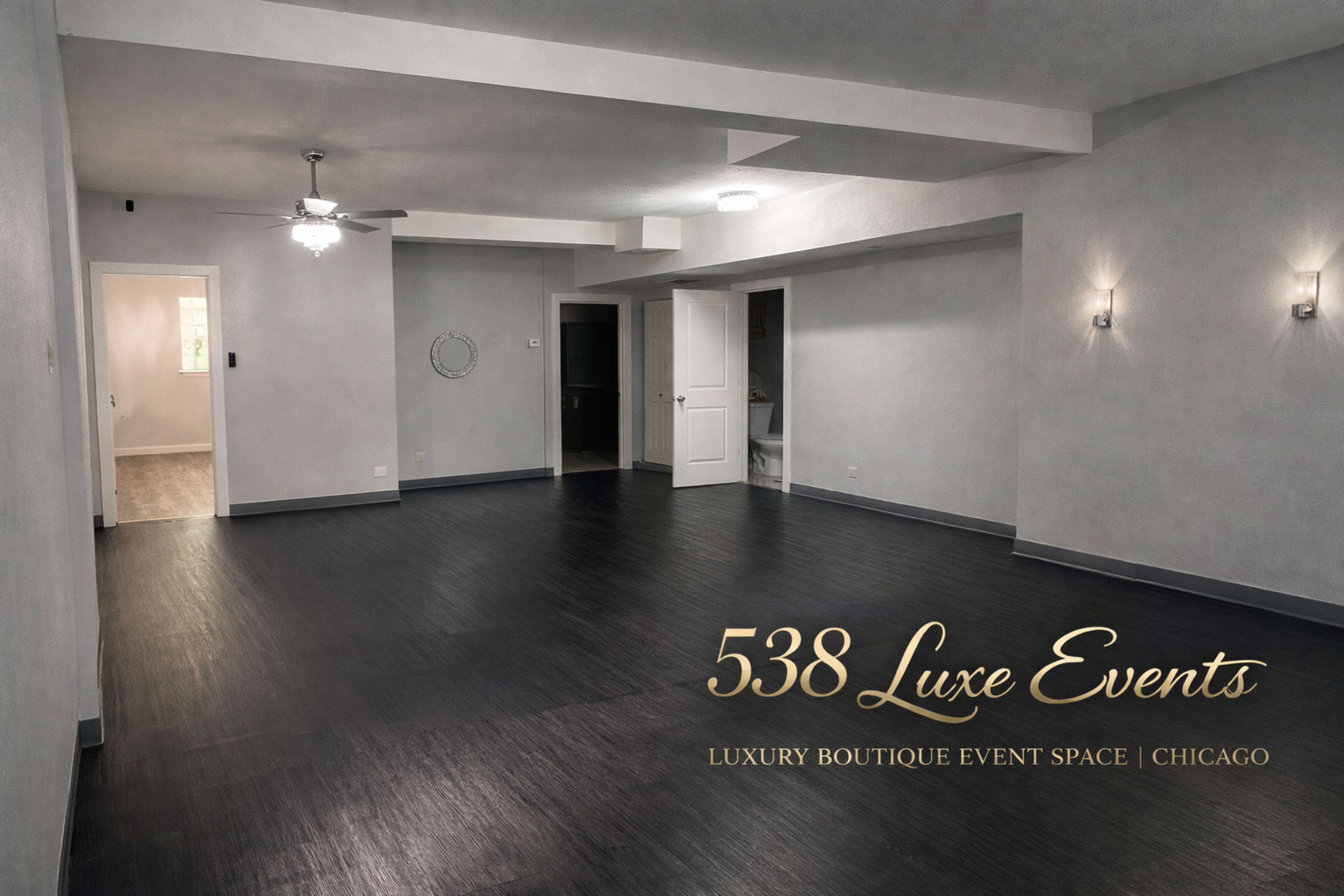 The image shows an empty event space with dark wood flooring, gray walls, and a ceiling fan, featuring a minimalistic and modern design.