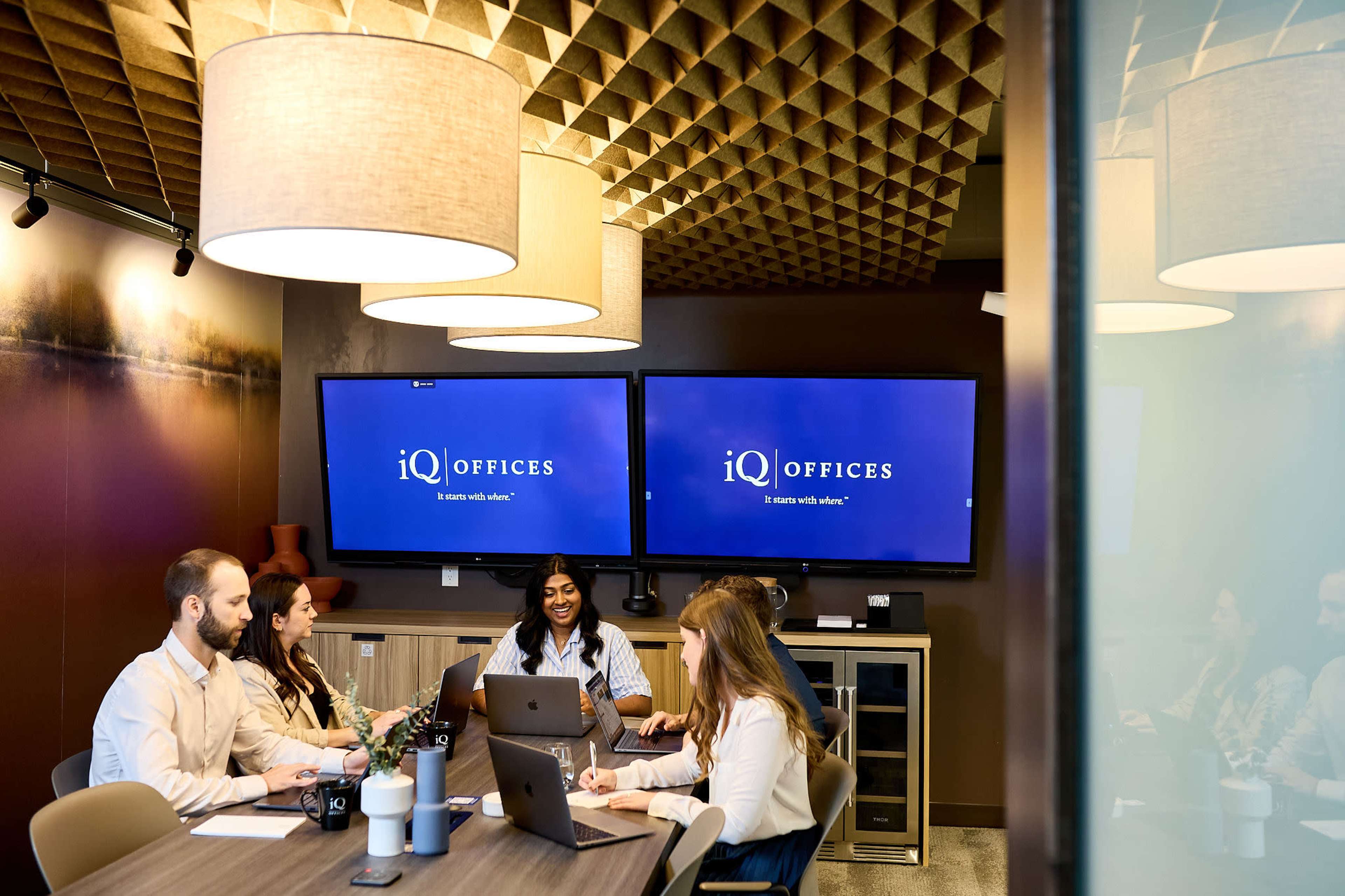 A group of four professionals are seated around a conference table in a modern meeting room, discussing ideas while using laptops, with two large screens displaying the logo "iQ OFFICES."