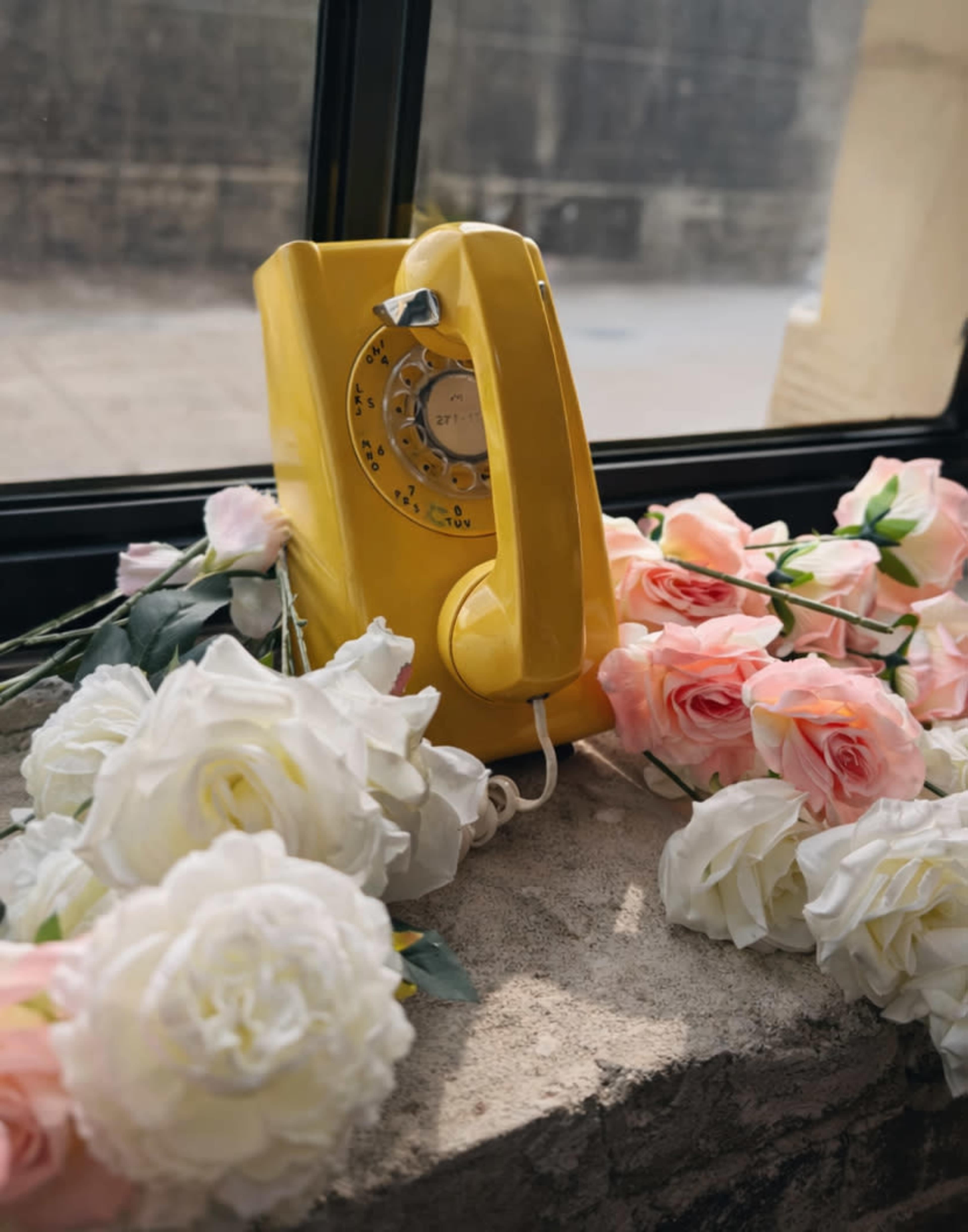 A yellow rotary phone is placed on a stone surface surrounded by white and pink artificial roses near a window.