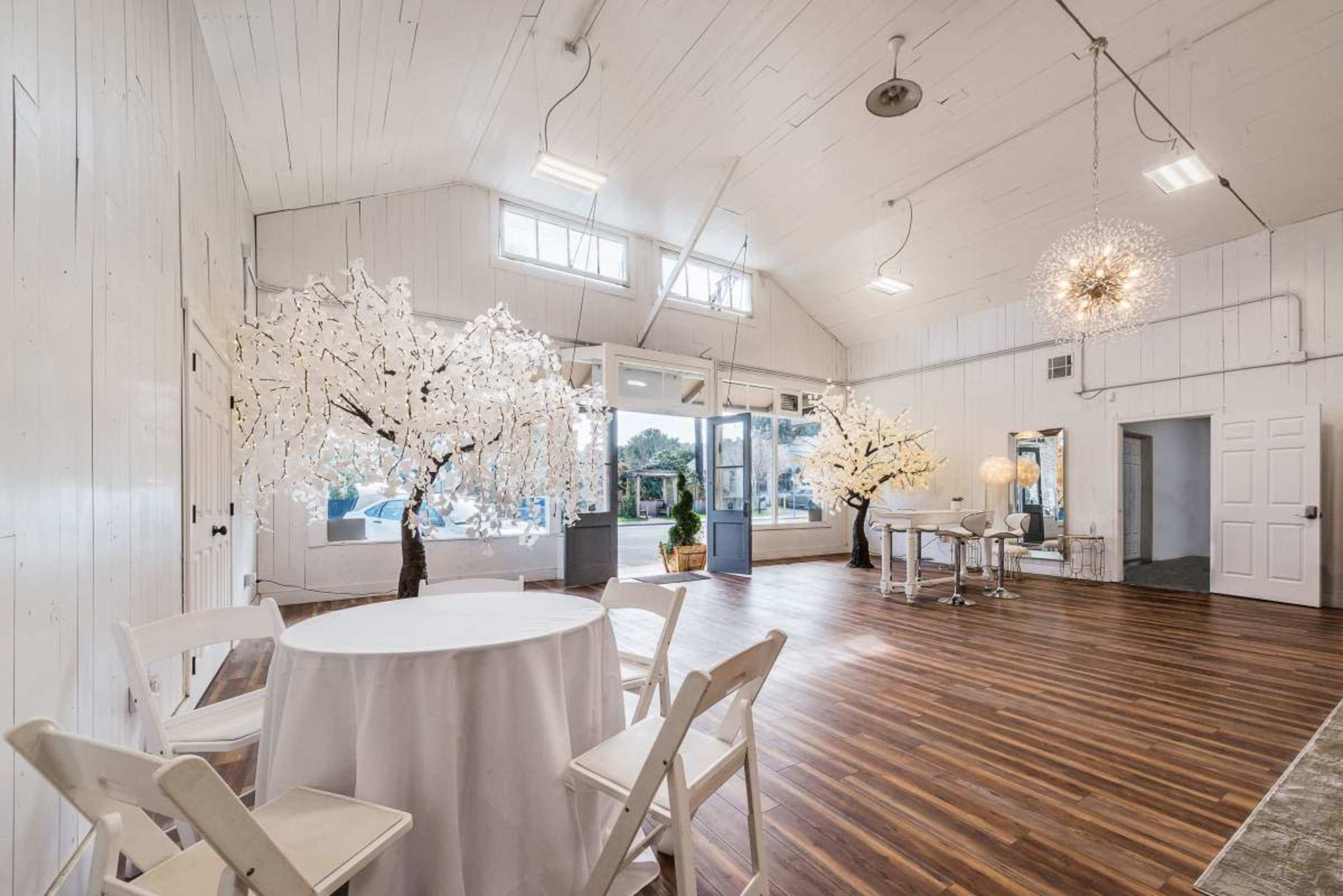 The image shows a spacious, well-lit interior featuring white walls, hardwood flooring, and decorative trees with white lights, along with a circular table and chairs arranged for an event.