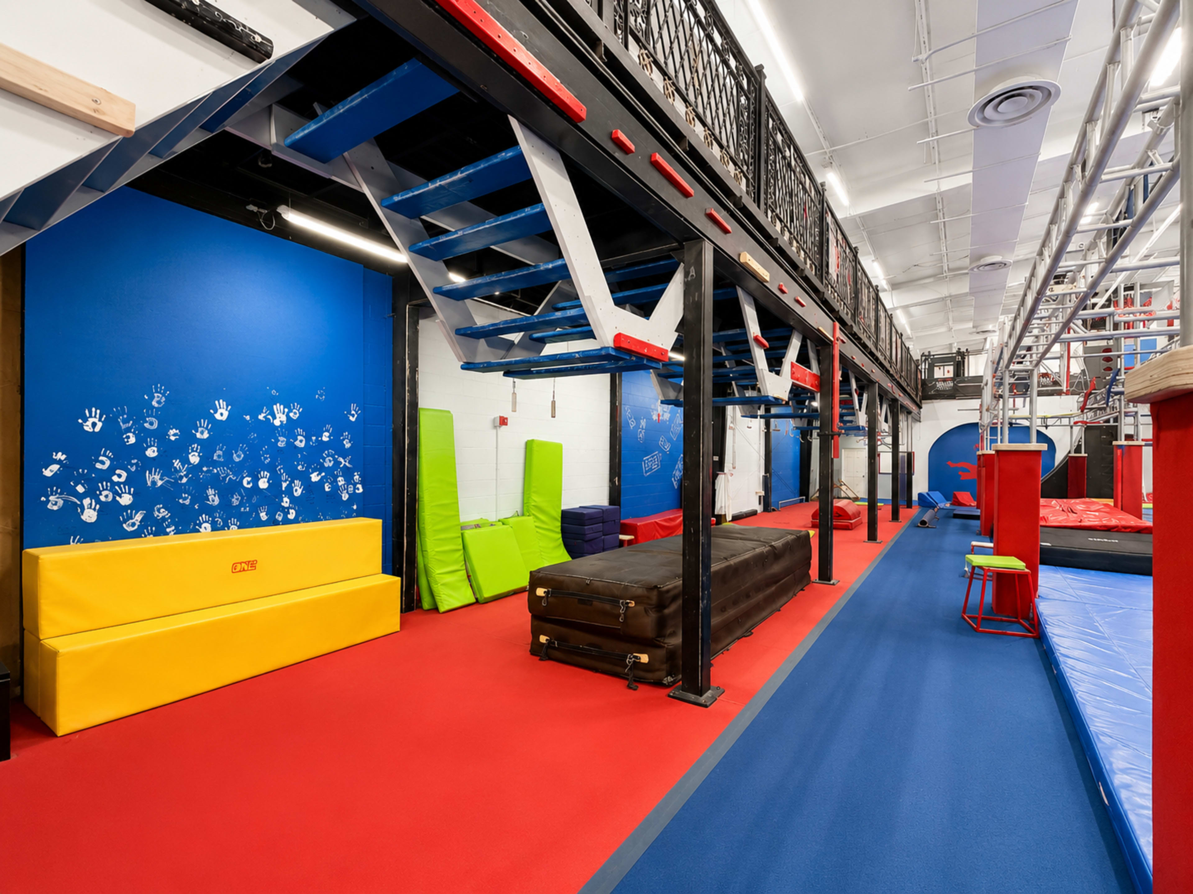 The image shows a brightly colored indoor gym space featuring various exercise equipment, including mats, a brown vaulting box, and colorful foam blocks.