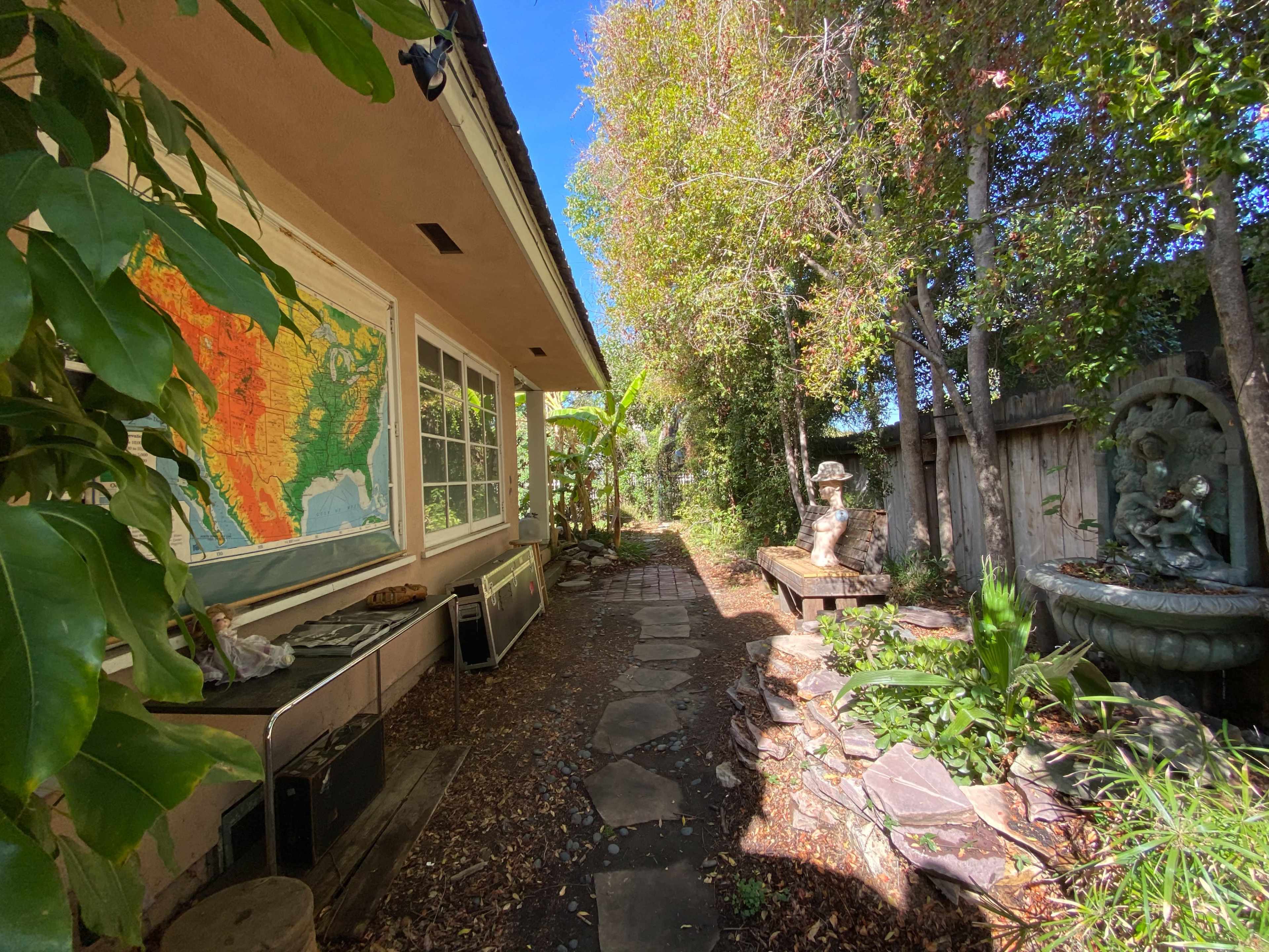 A narrow pathway lined with stones, flanked by greenery and a map on the wall of a building, leading to a tranquil garden area with a fountain.