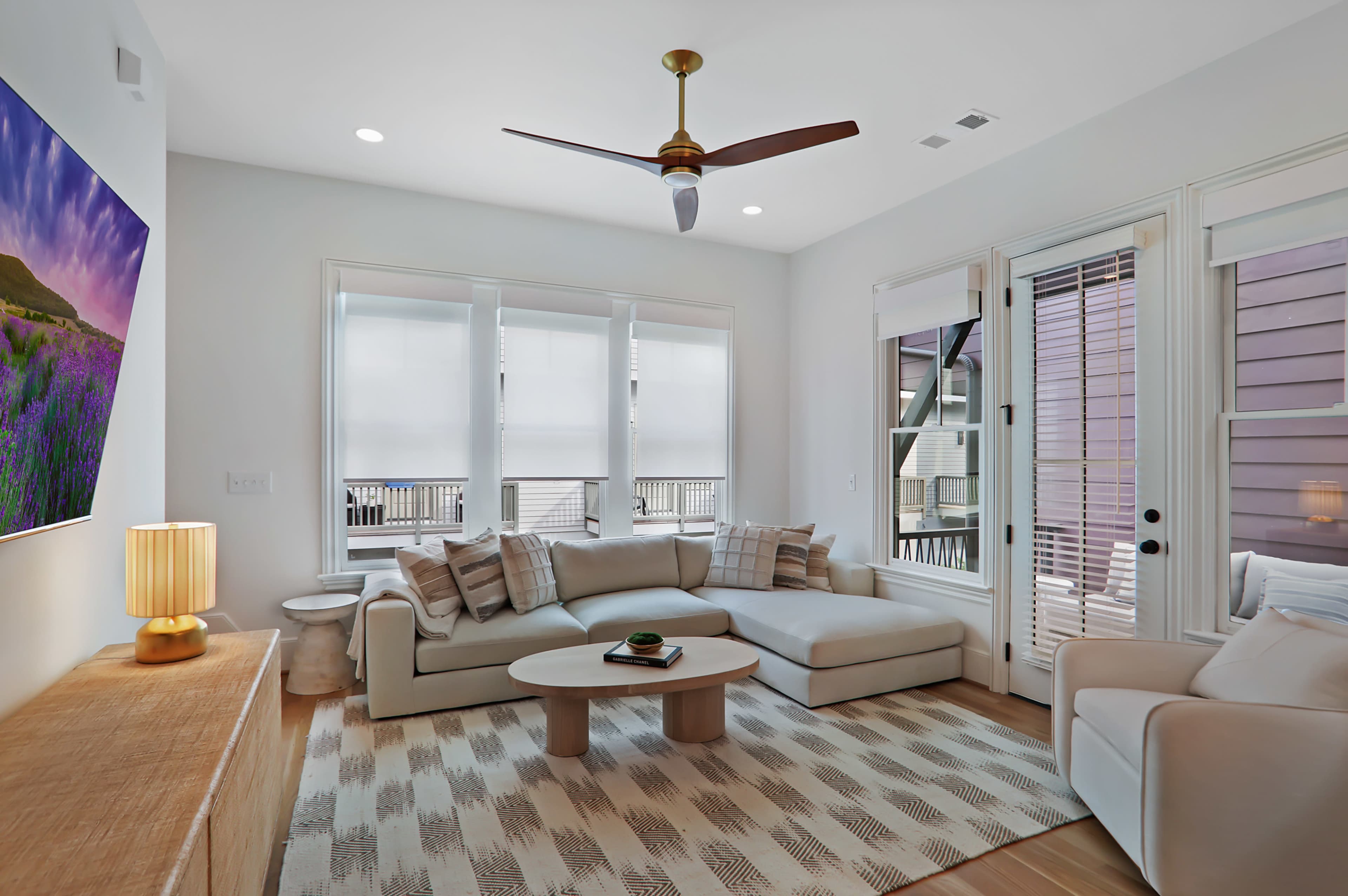 The living room features a spacious sectional sofa, a round coffee table, and a television mounted on the wall, with large windows letting in natural light.