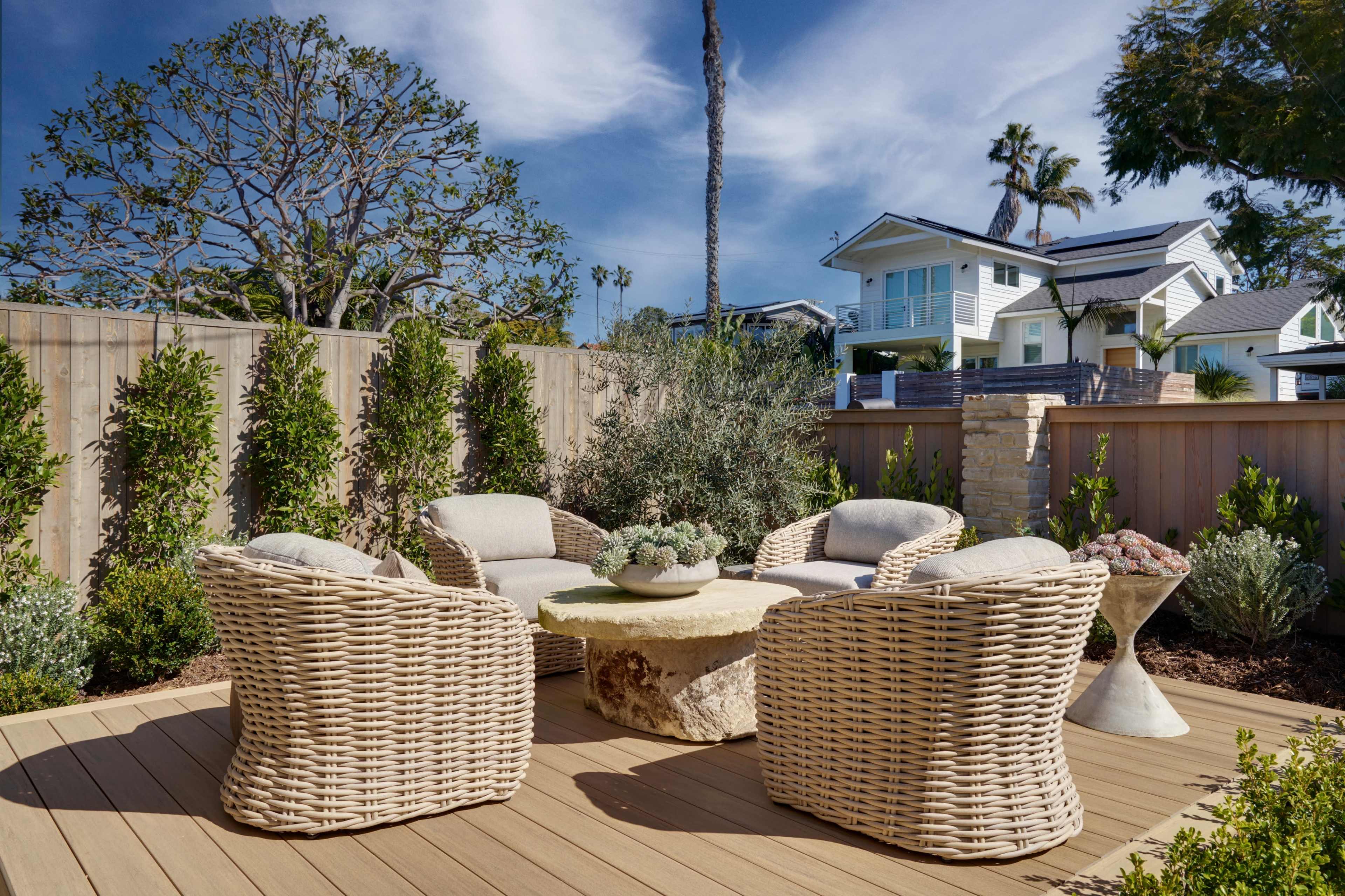 A seating area with wicker chairs and a stone table is set on a wooden deck surrounded by lush greenery and a wooden fence.