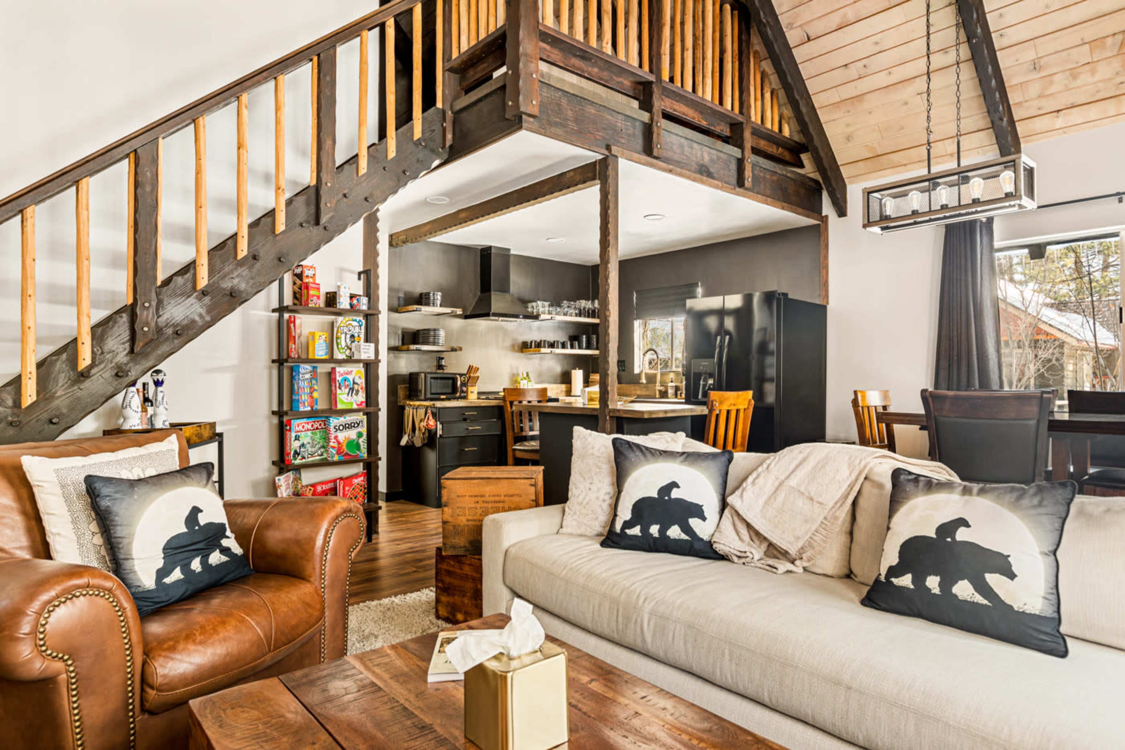 A cozy living space features a mixture of leather and fabric sofas, a wooden table, and a staircase leading to an upper level, with a kitchen and dining area visible in the background.