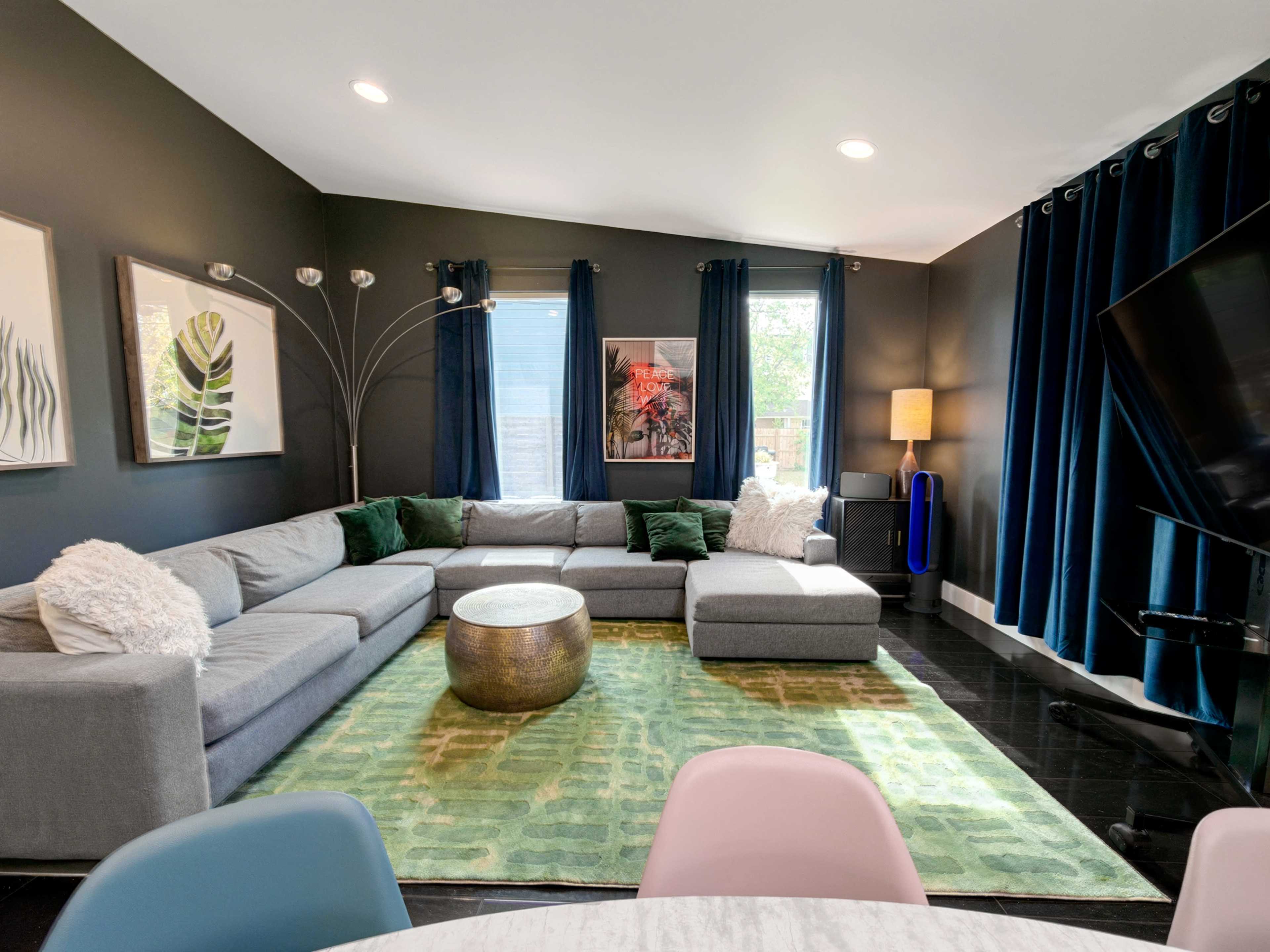The image shows a modern living room with a grey sectional sofa, a round metallic coffee table, green accents, and large windows covered with dark curtains.