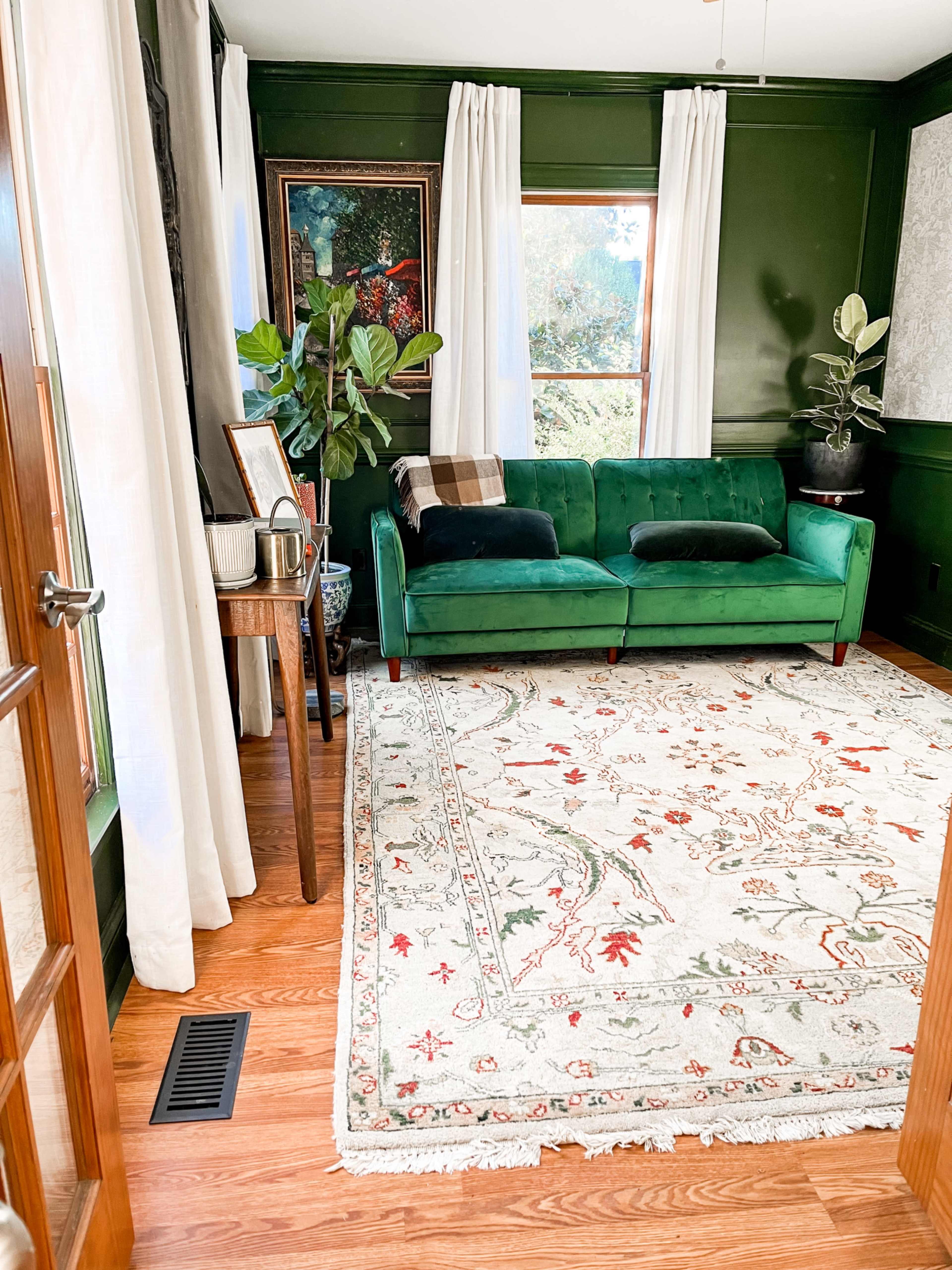 The room features a green velvet sofa facing a large window, surrounded by houseplants and a decorative rug on the wooden floor.