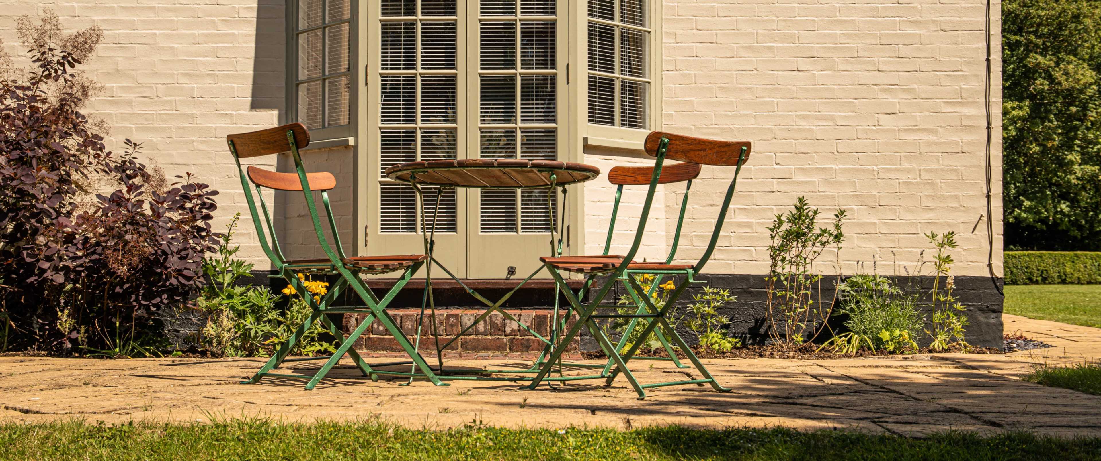A small round table with two folding chairs sits on a stone patio beside a house with a light-colored brick wall and large windows.