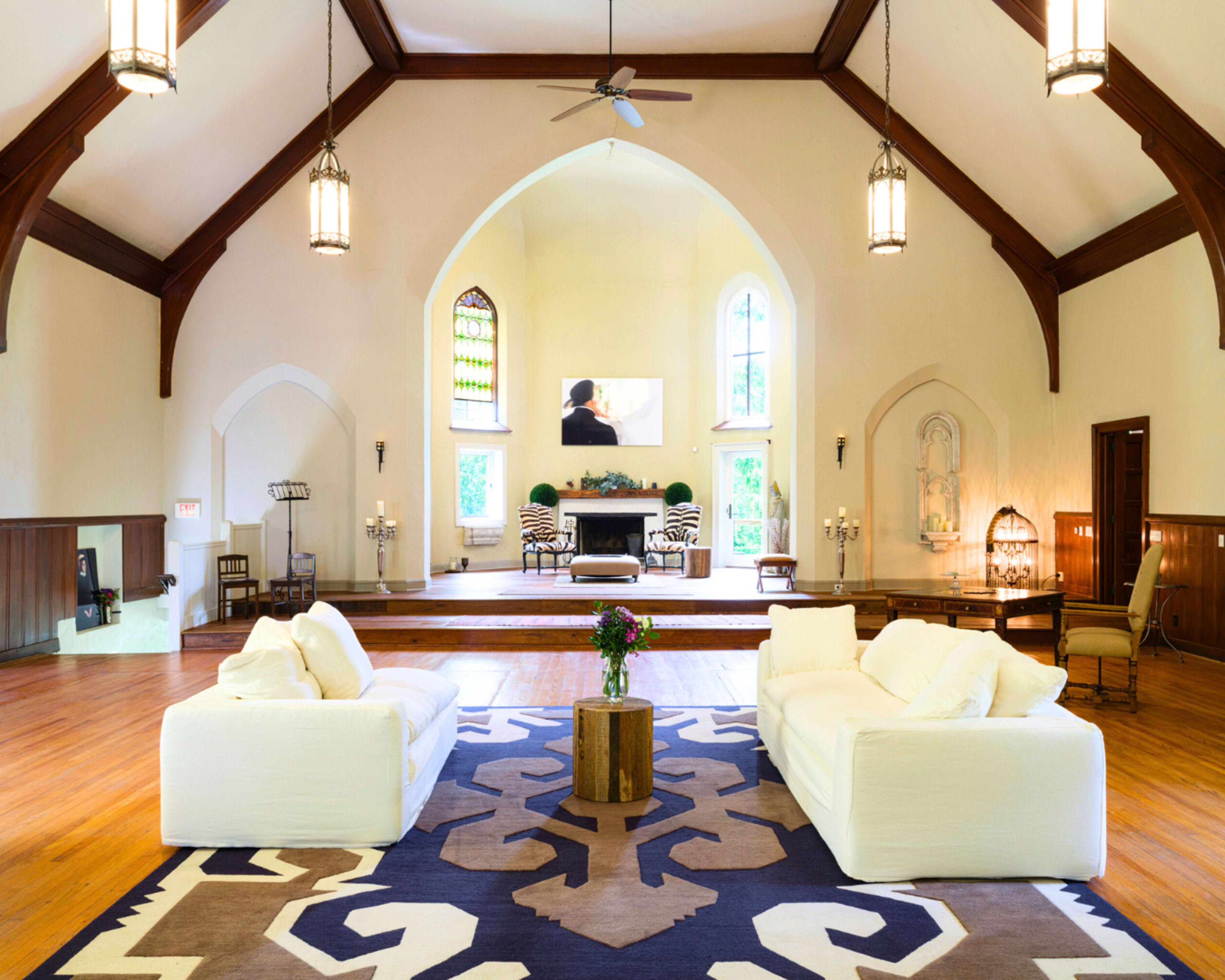 The image shows a spacious, elegantly designed living room with high wooden ceilings, large windows, and two white sofas facing a stage-like area with a fireplace.