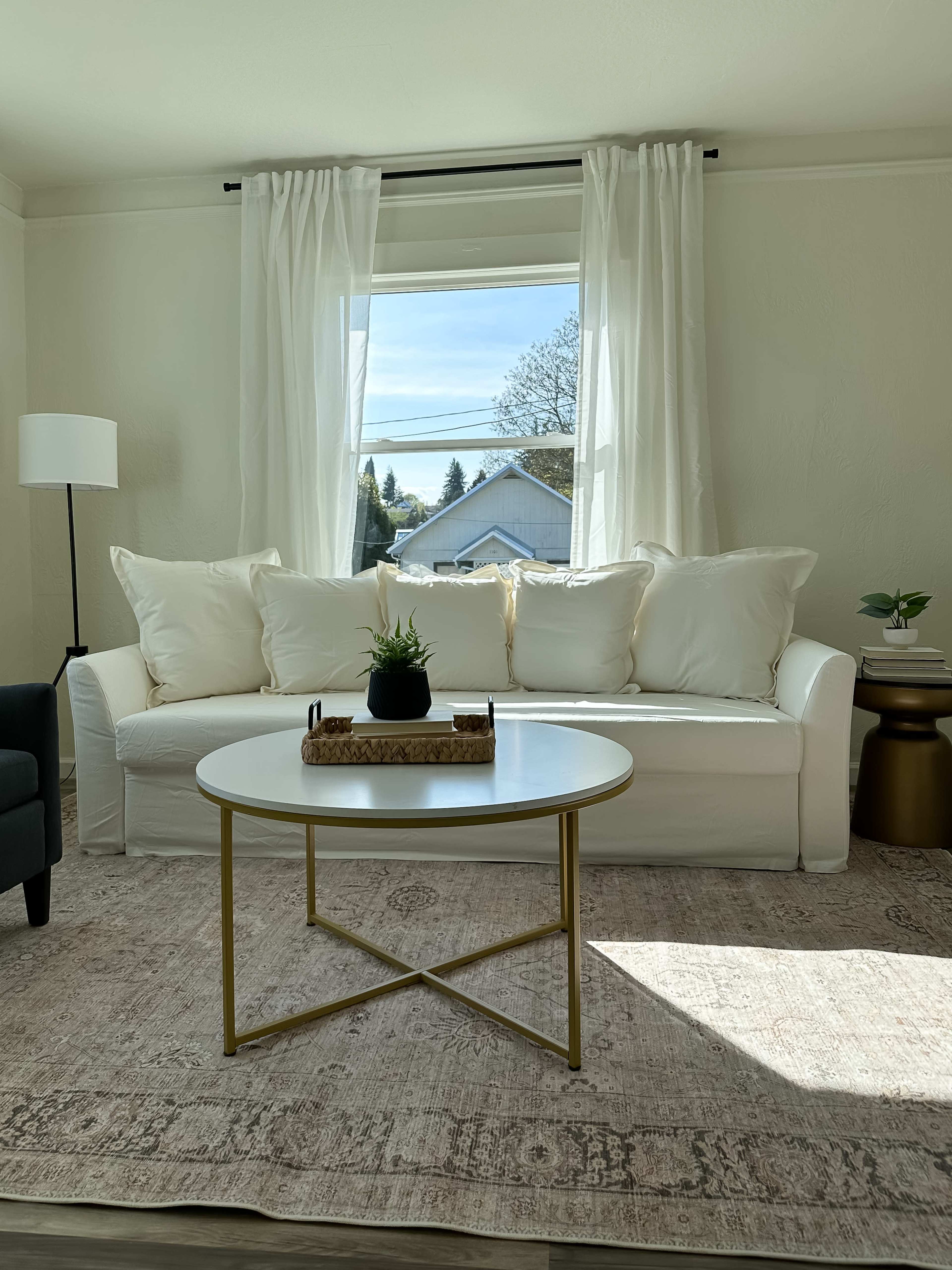 A bright living room features a white sofa with decorative pillows, a round coffee table, and large windows letting in natural light.