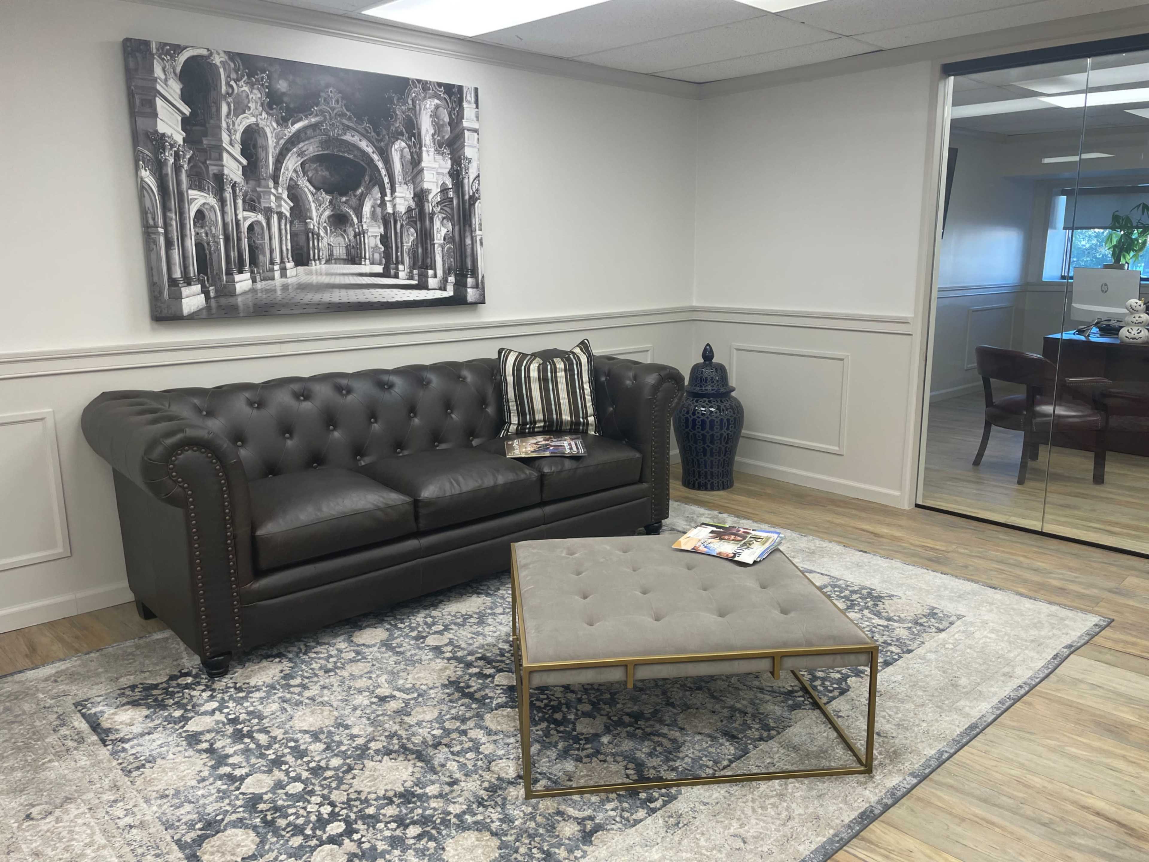 A modern office lounge area featuring a dark leather tufted sofa, a coffee table, and a large black and white photograph on the wall.