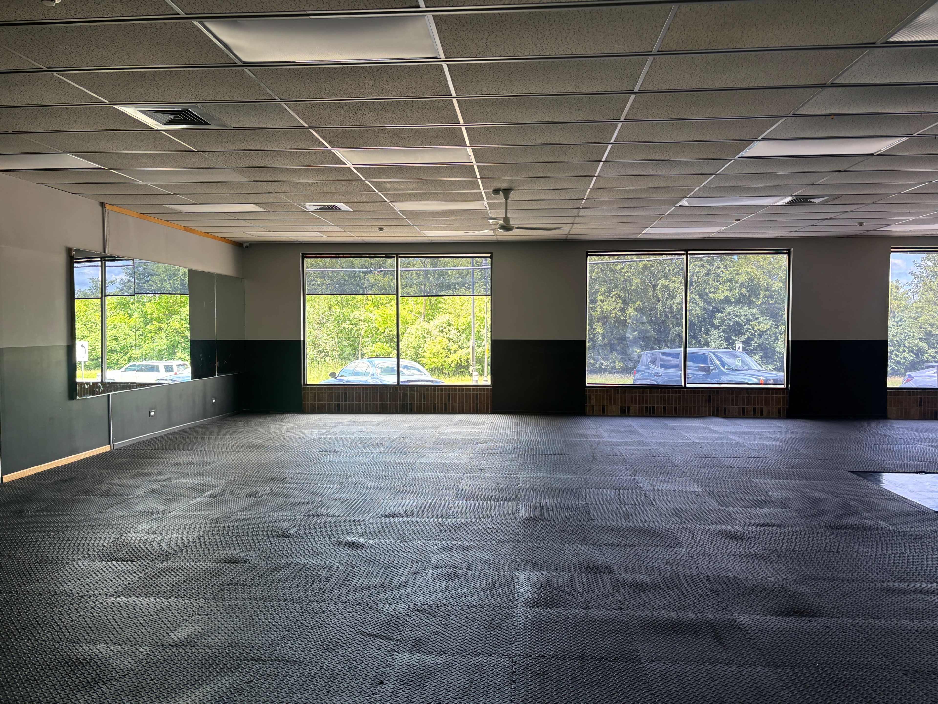 The image shows an empty, spacious room with black rubber flooring, large windows allowing natural light, and mirrors on one wall.