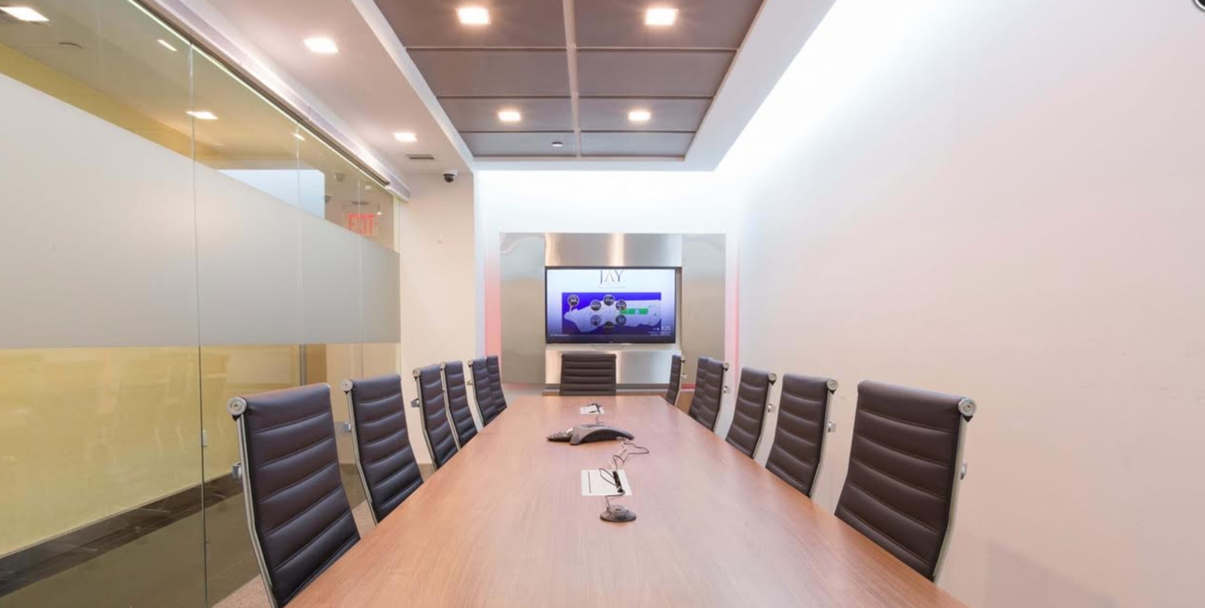 A long conference table with black leather chairs is arranged in a modern meeting room featuring a large screen at one end.